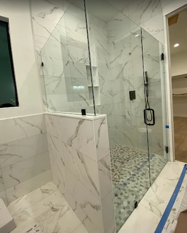 A walk-in shower with marble-look wall tiles, a pebble-stone floor, a glass door, and a built-in shelf.