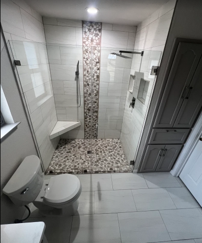 A modern bathroom features a glass-enclosed shower with tiled walls, pebble flooring, a built-in corner seat, and a toilet.