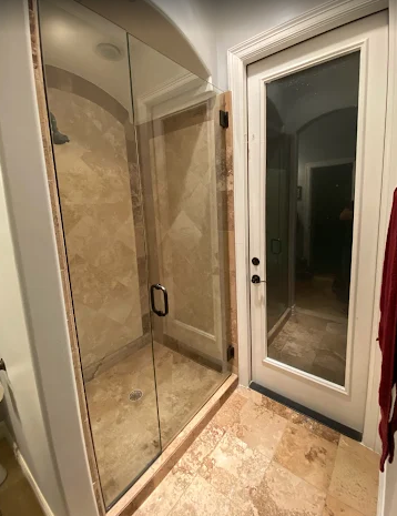 A tiled shower with a glass door next to a white door with a glass pane in a bathroom with beige stone floors.