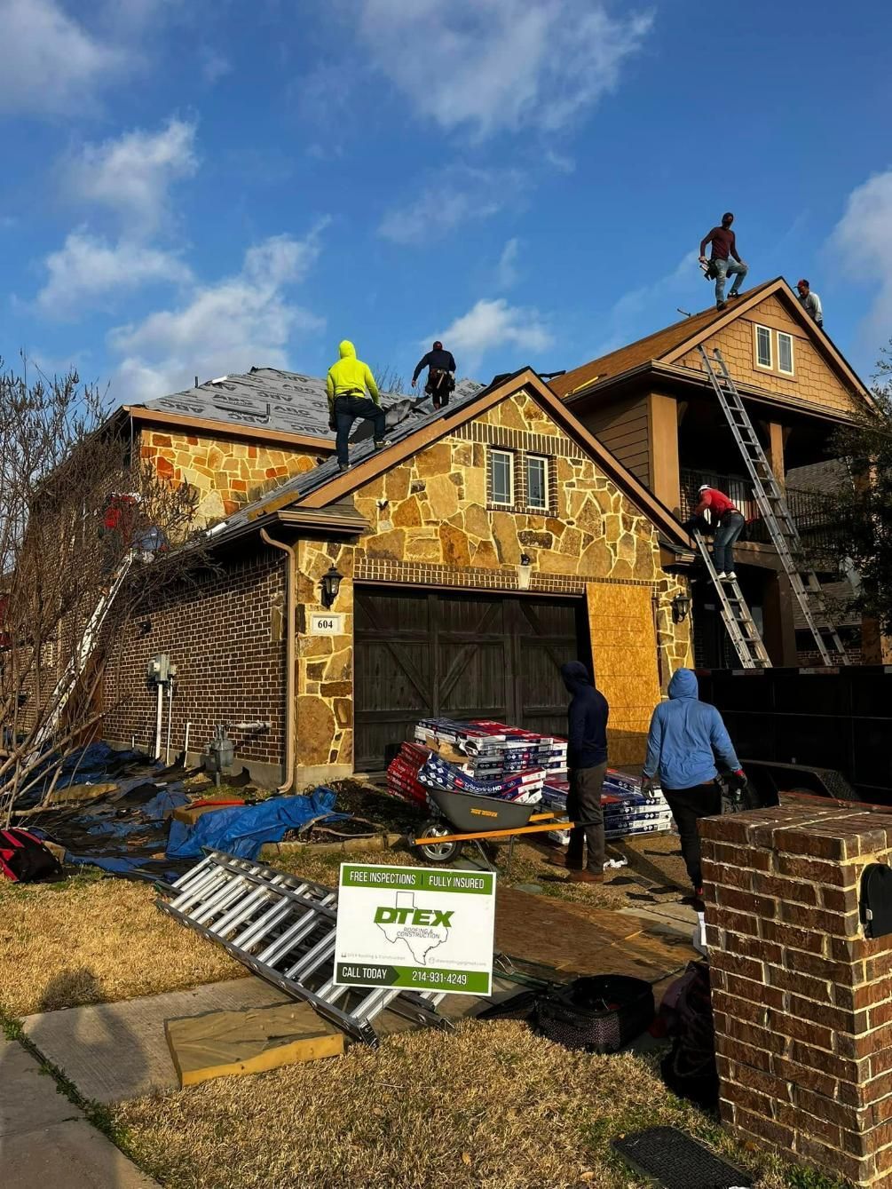 Roofers working on a residential roof, some on ladders, with materials and tarps on the ground.