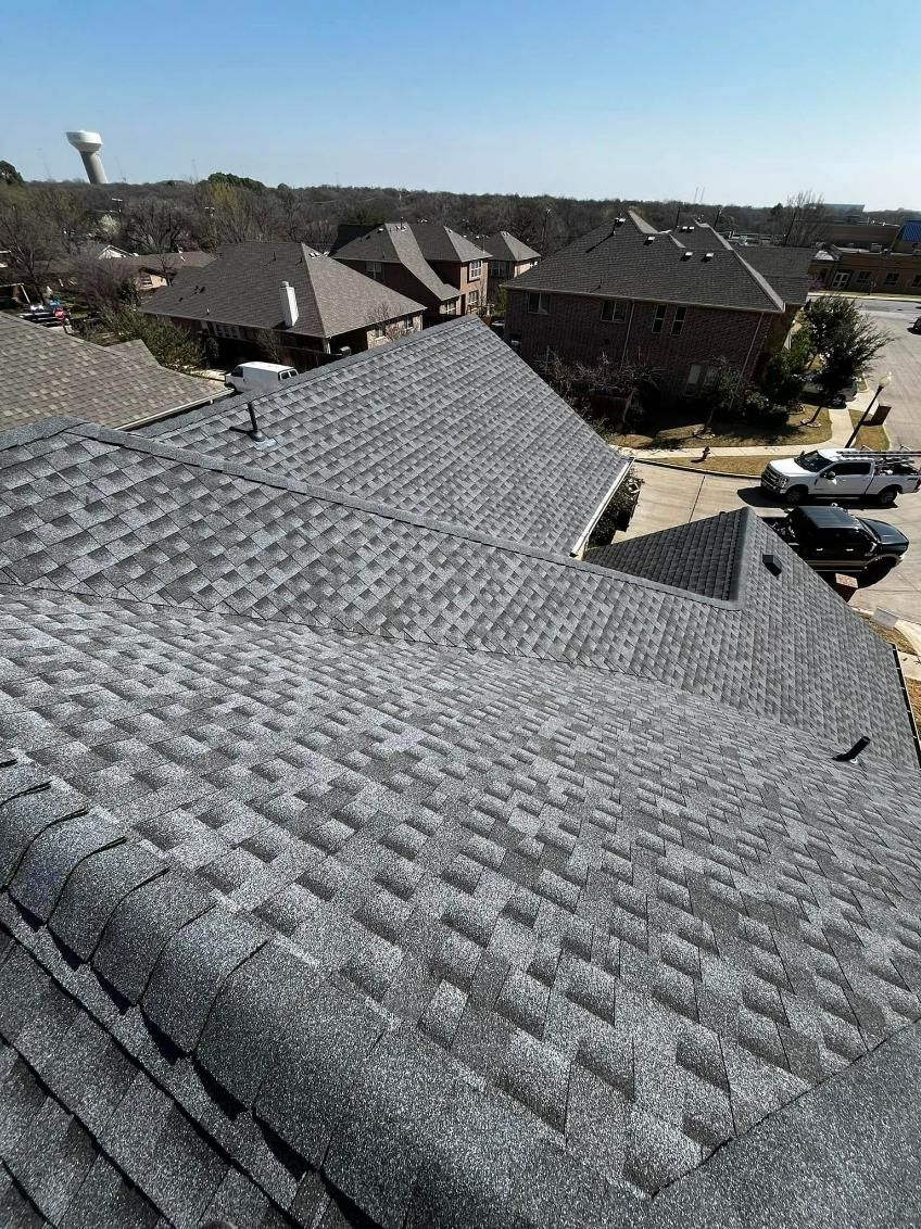 Gray asphalt shingle roof on a sunny day, with a neighborhood in the background.