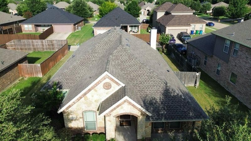 Aerial view of a suburban house with a brown shingle roof, surrounded by other homes and green lawns.