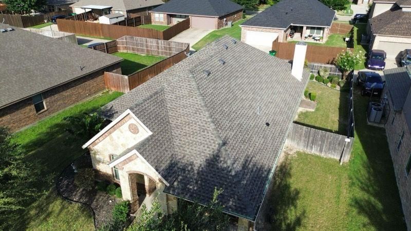 Aerial view of a suburban house with a brown roof and attached garage, surrounded by green lawns and brown fences.
