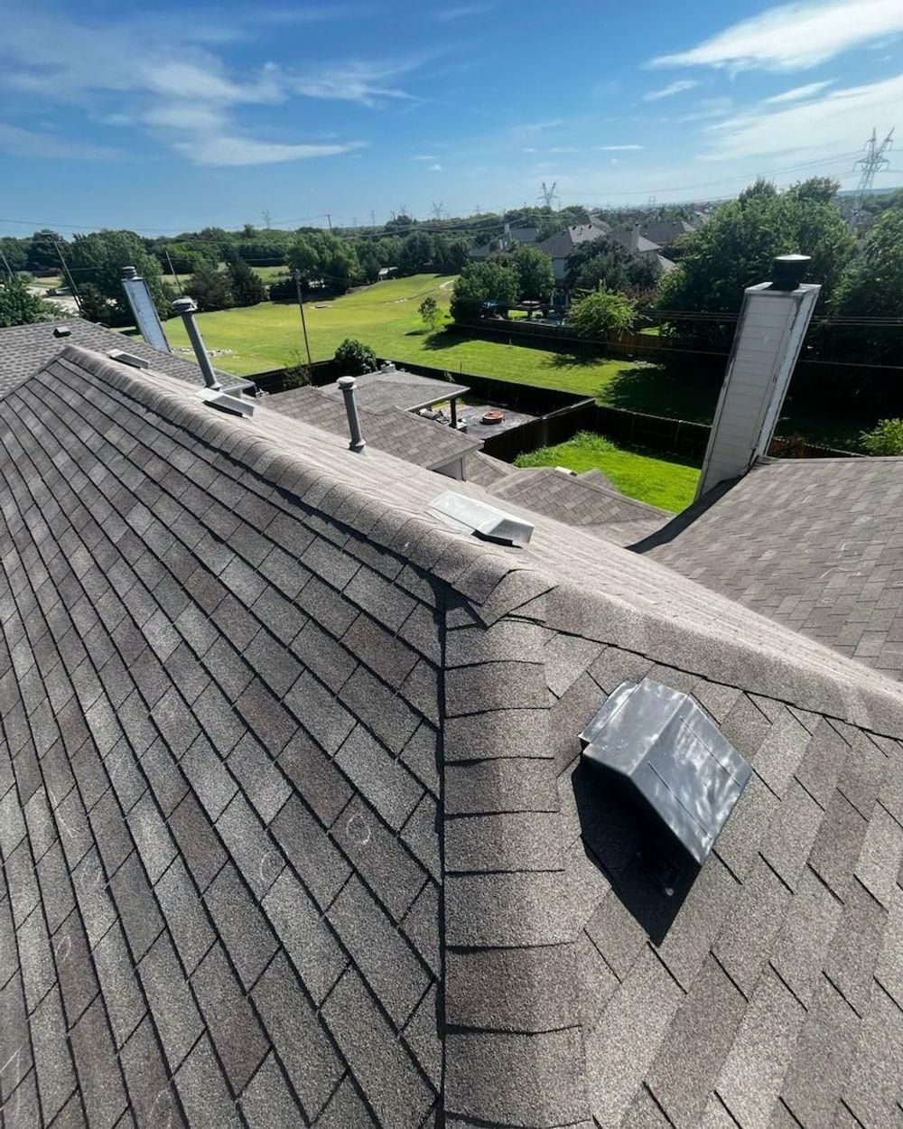 View of a shingled roof with chimneys and vents against a blue sky, overlooking a green landscape.