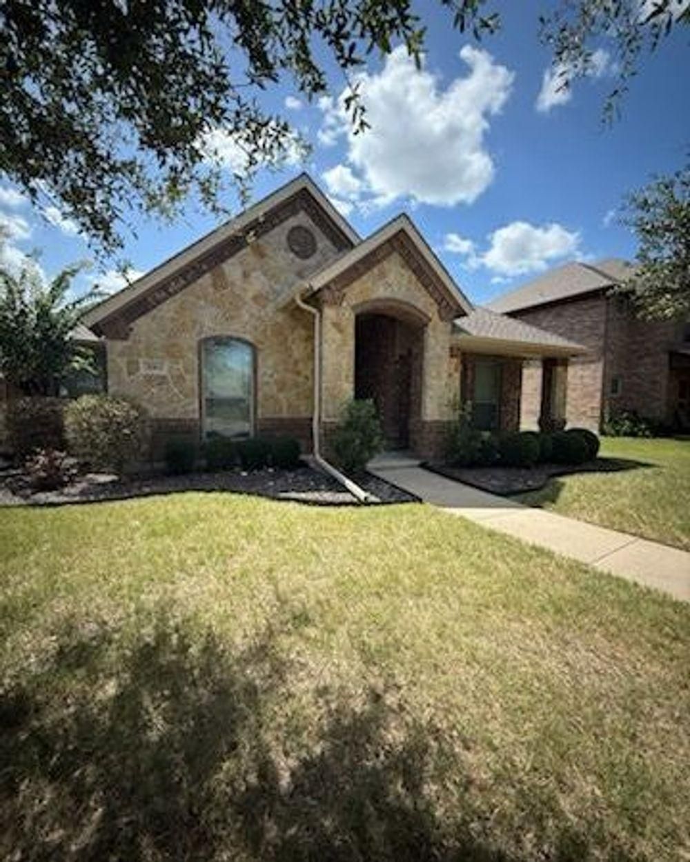 Stone-façade house with a covered entrance on a sunny day with clouds. Green lawn and trees in the front yard.