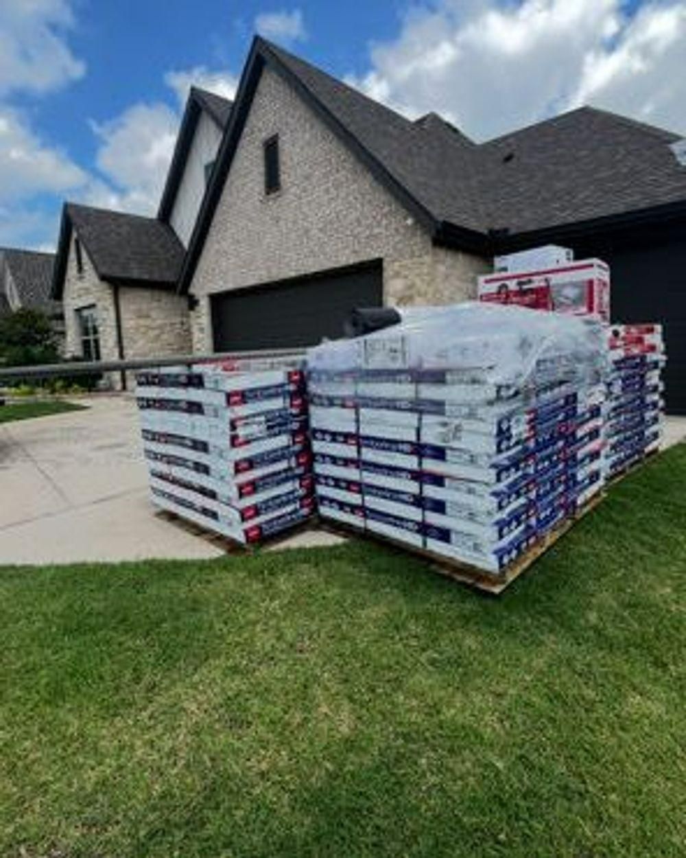 Pallets of roofing materials stacked on a grassy lawn in front of a house.