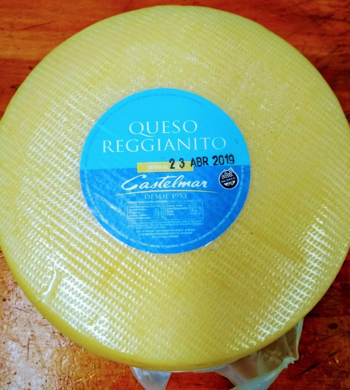 Rueda redonda de queso Queso Reggianito con etiqueta azul sobre una superficie de madera.