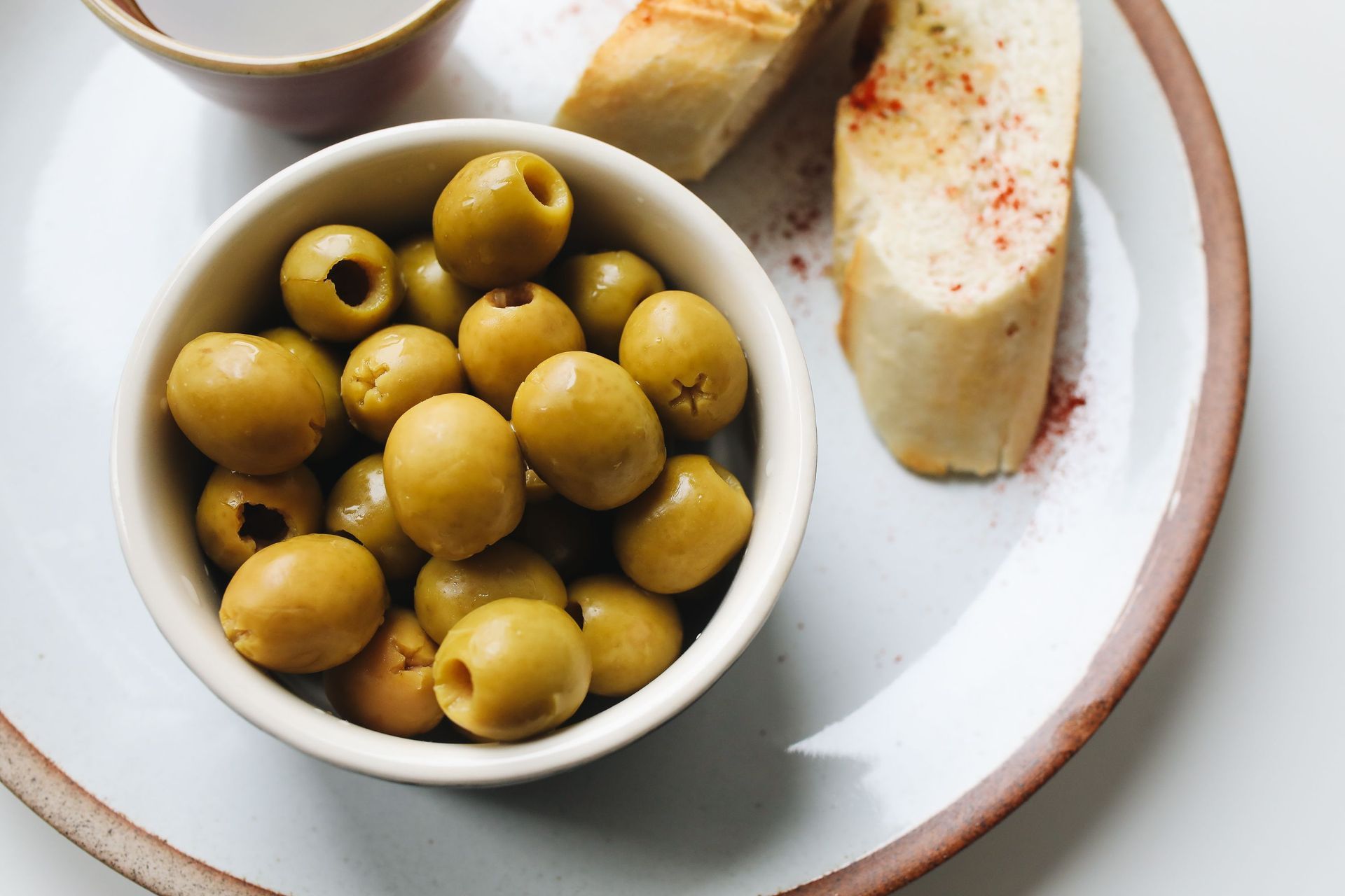 Bol de aceitunas verdes y pan rebanado en un plato blanco con salsa para mojar.