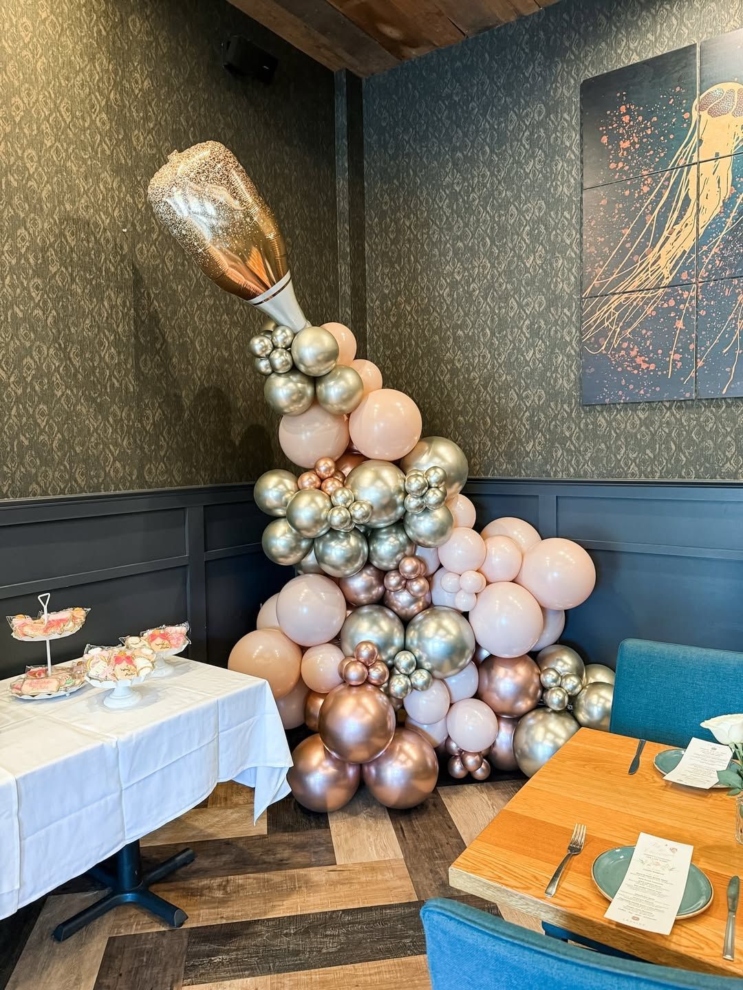 A champagne bottle foil balloon with a cascade of pink, silver, and gold balloons in a corner by a restaurant table.