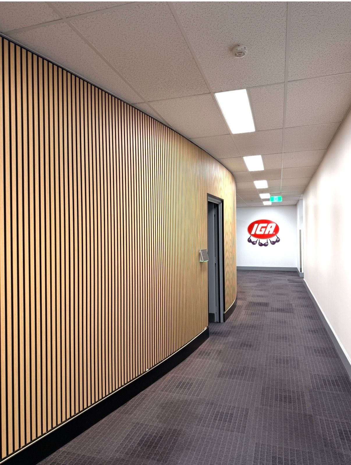 Empty office hallway with wood-slat wall, white walls, gray carpet, and ceiling lights leading to a logo at the end