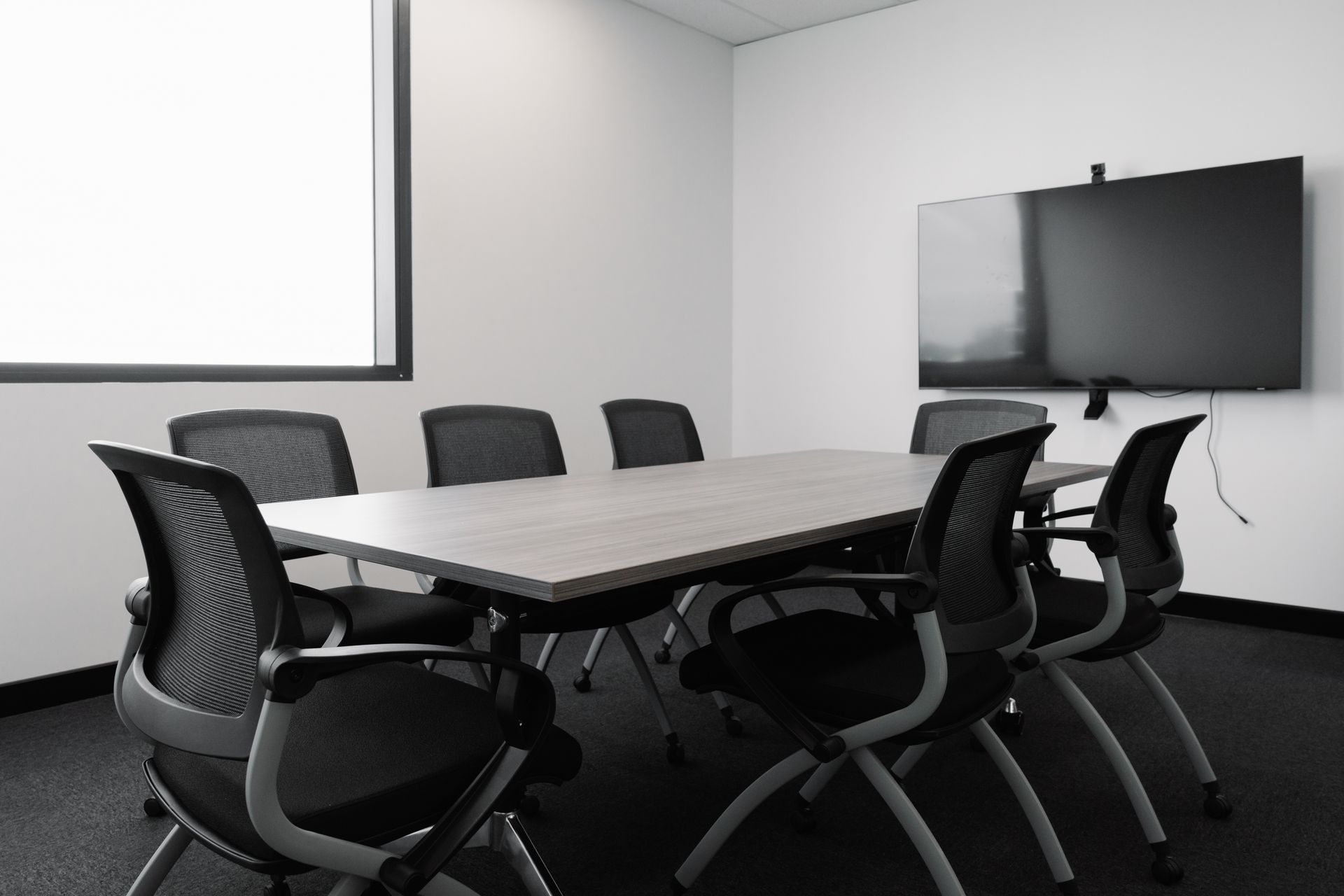 A conference room with a rectangular table, eight chairs, a large screen, and a window.