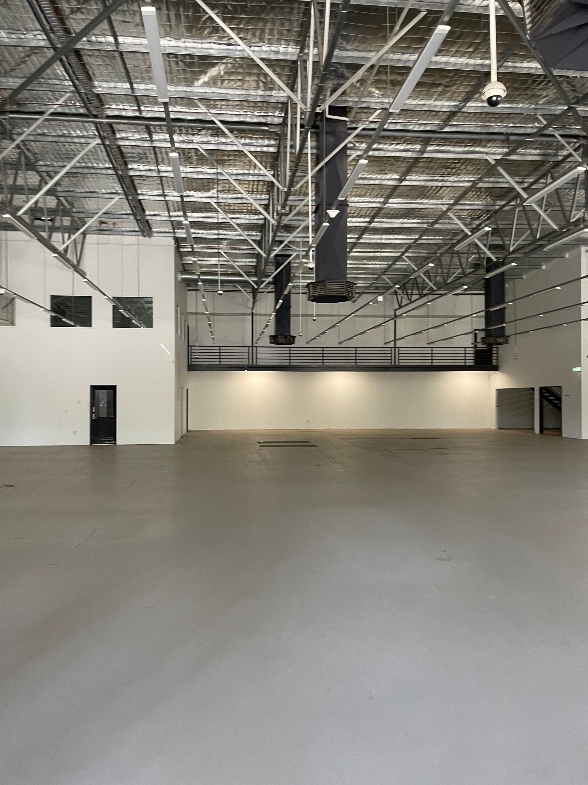 Empty industrial space with high ceilings, metal beams, and gray floor. White walls, black doors, and overhead vents.