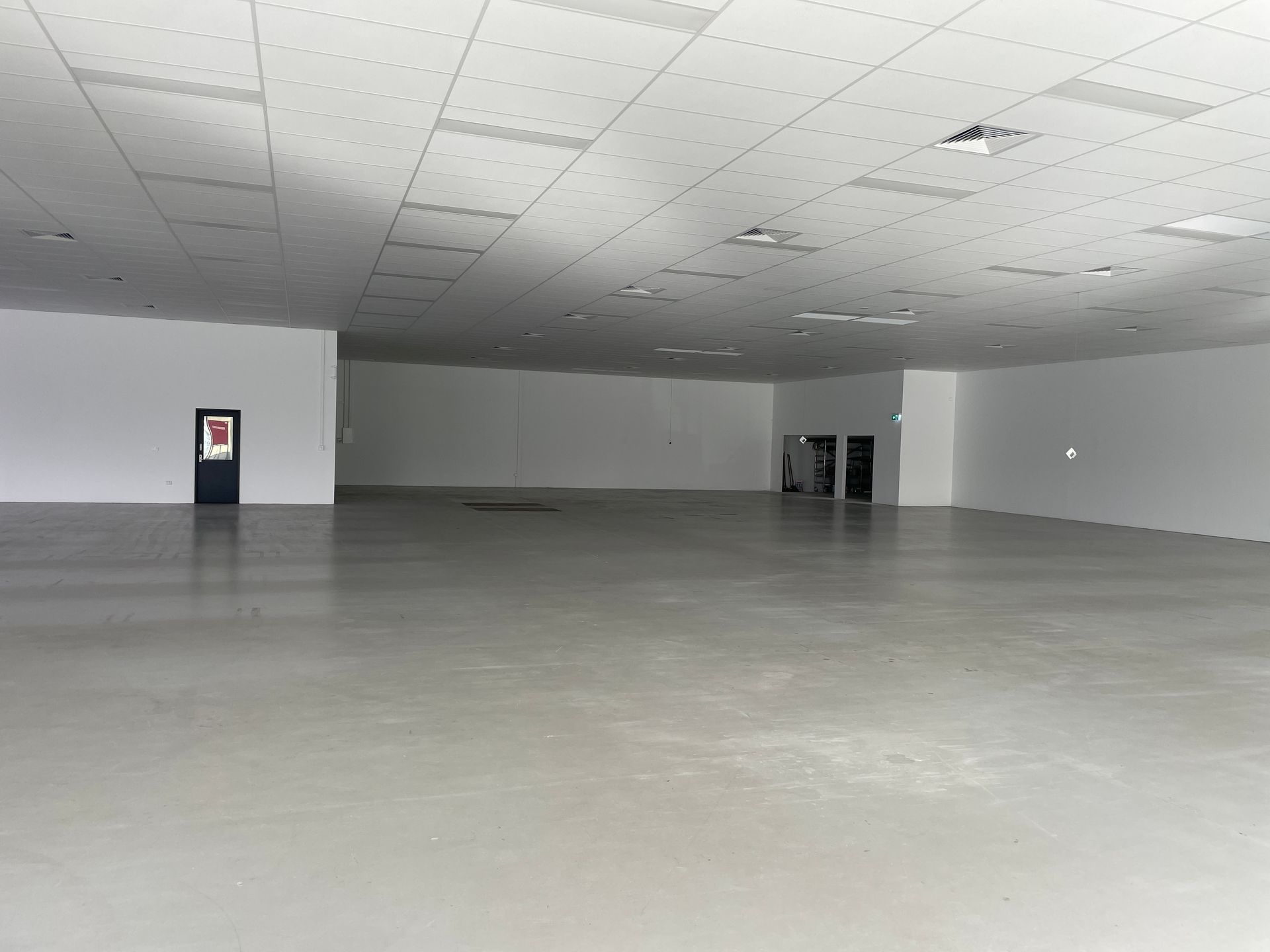 Empty, spacious room with white walls, gray floor, and a suspended ceiling with lights. Dark doorway on the left.