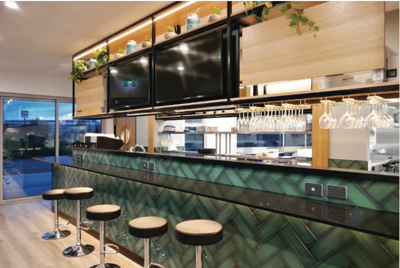 Green tiled bar with stools, TVs, and hanging glasses, featuring an open kitchen and outdoor view.