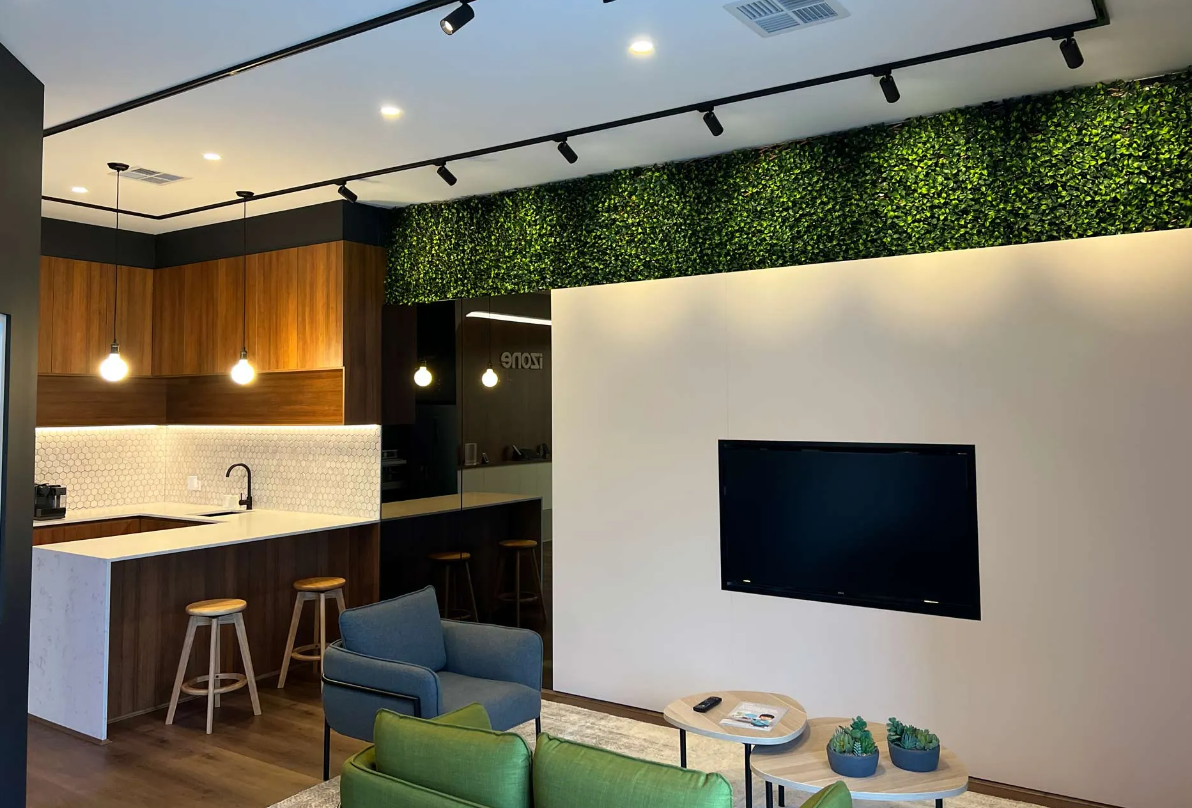 Modern living room with kitchen. Brown cabinets, white counters, green wall accent, TV, and track lighting.