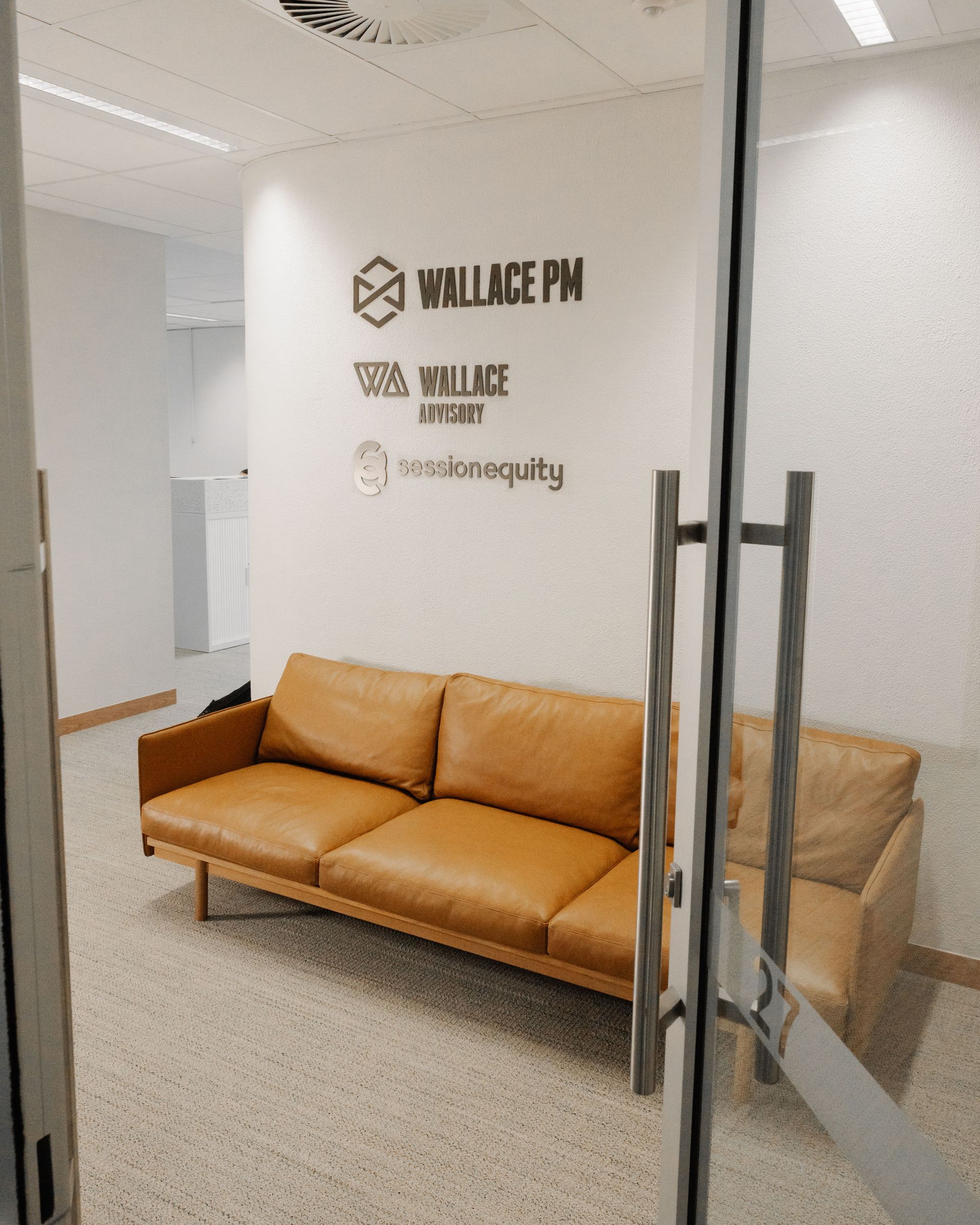 A modern office waiting area. Tan leather couch, signage on white wall, through glass door.