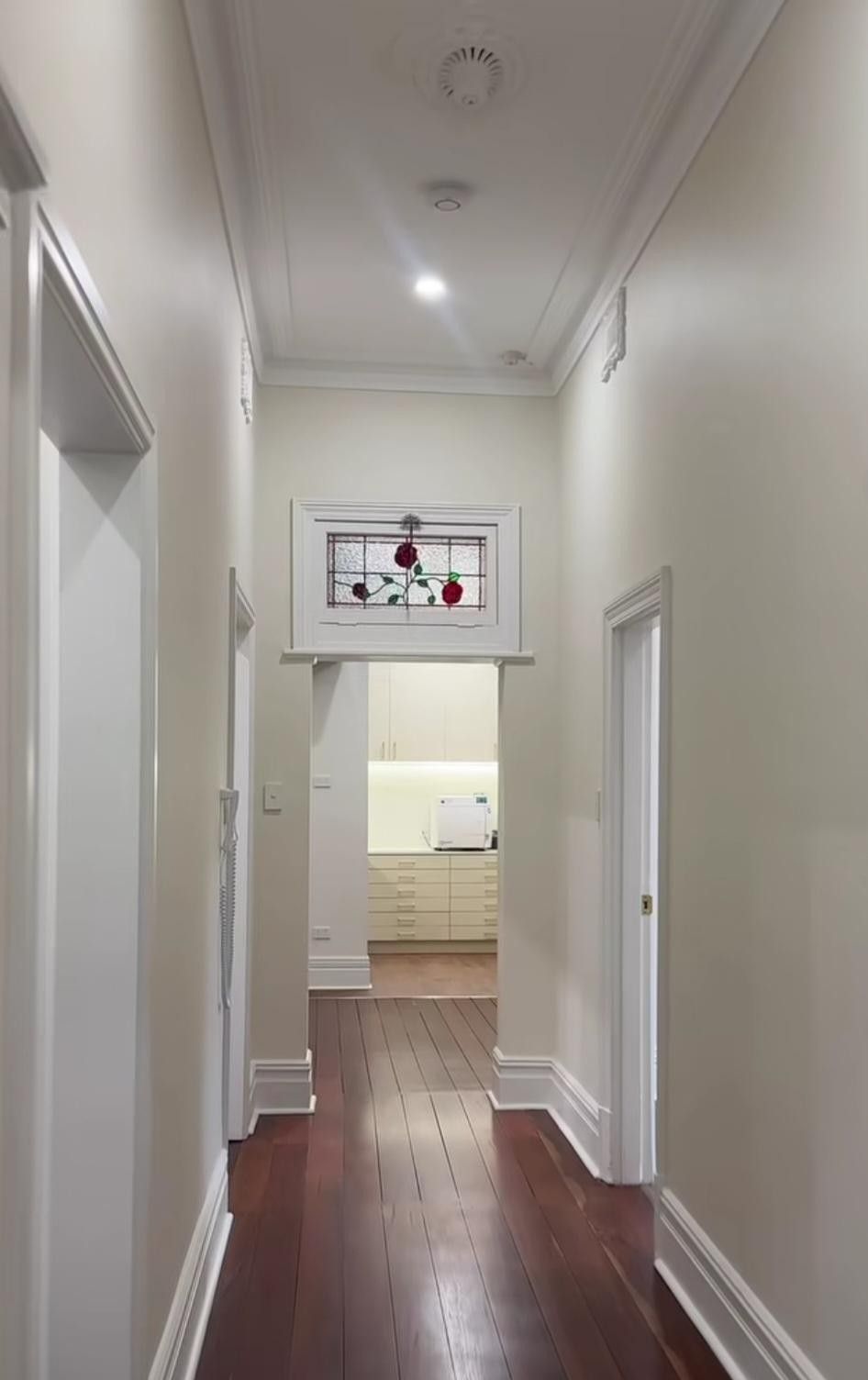Narrow white hallway with hardwood floors, decorative glass transom, and a room visible at the end.