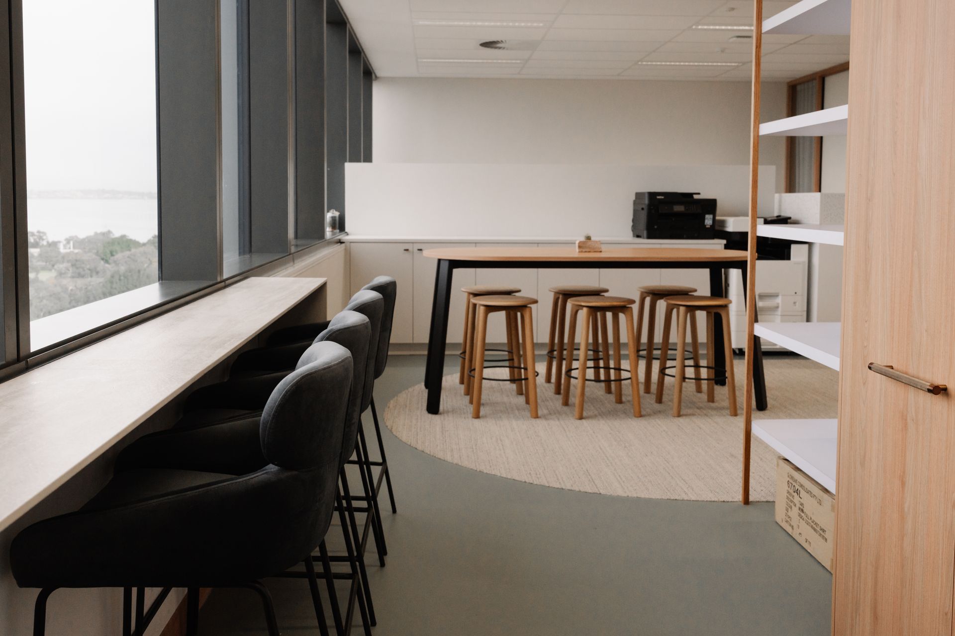 Modern office space with window seating and a central table with stools, plus shelving and a printer.