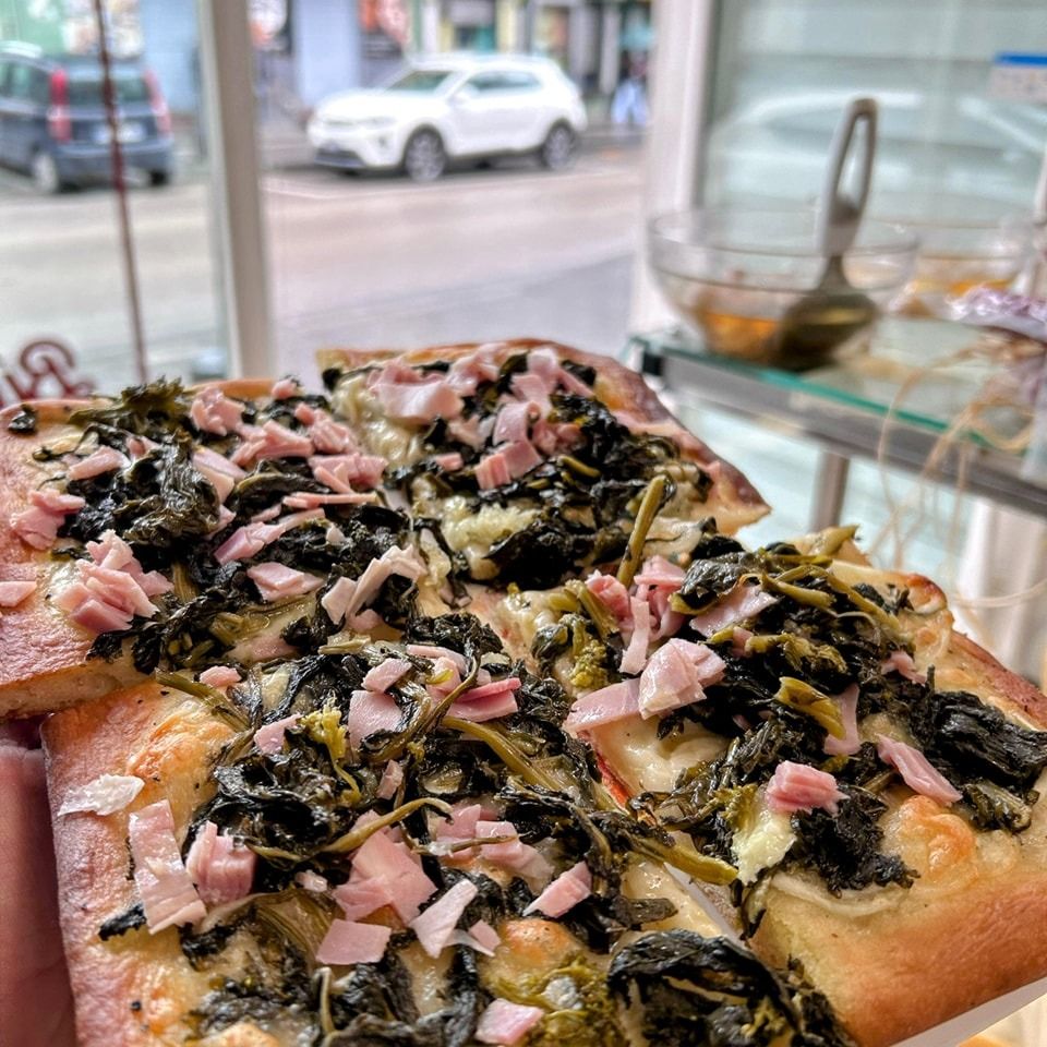 Pizza quadrata con verdure e prosciutto, servita vicino a una finestra con vista sulla città.