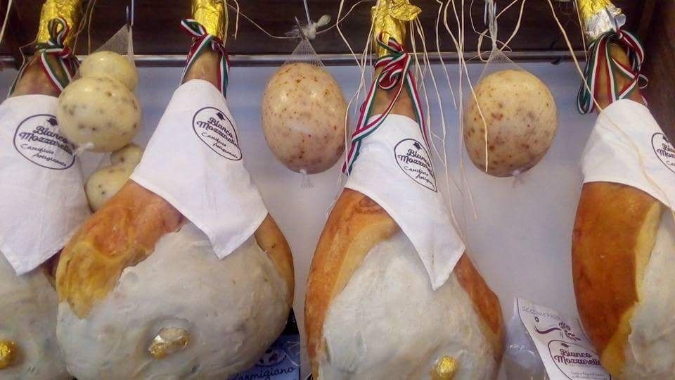 Cosce di prosciutto stagionato appese insieme a formaggi rotondi, in un'esposizione di negozio.