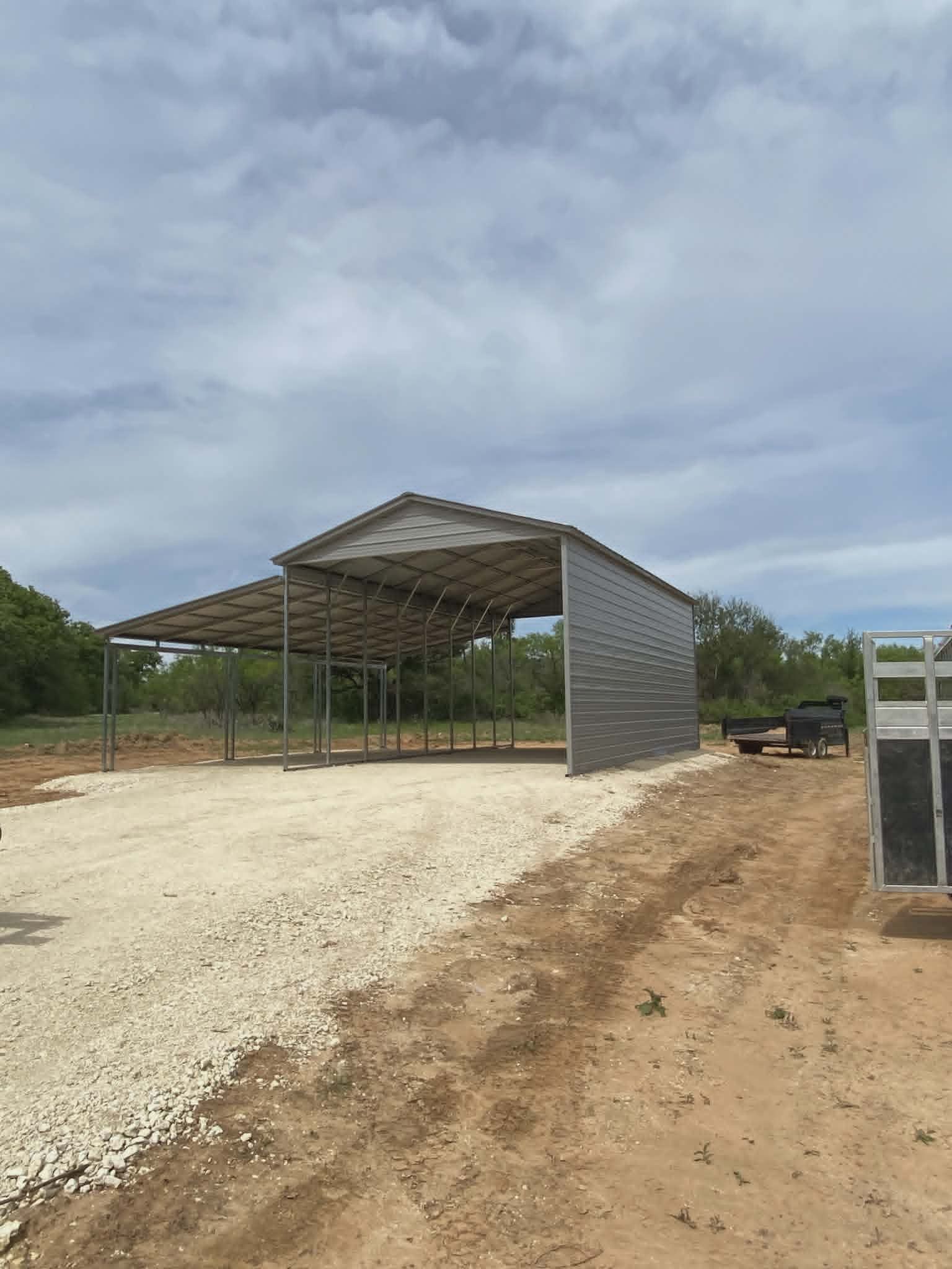 Open-sided metal shed under construction on a gravel lot with a cloudy sky overhead