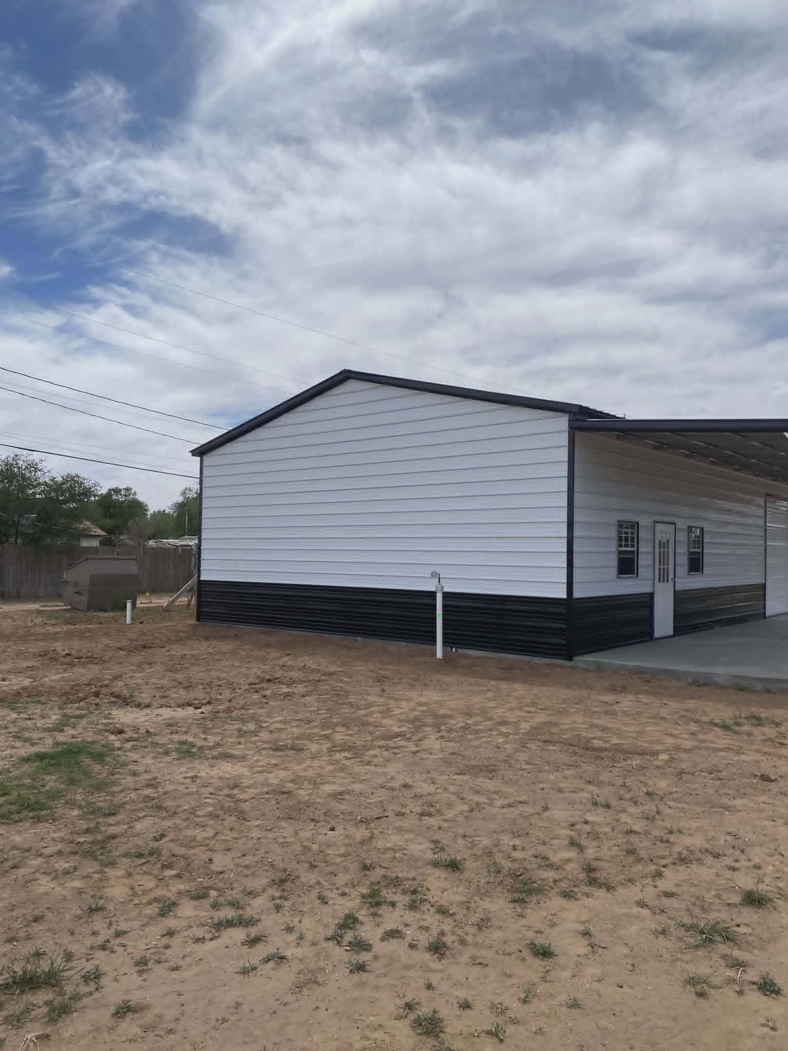 Small white house with black trim and porch, set on a bare dirt lot under a cloudy sky