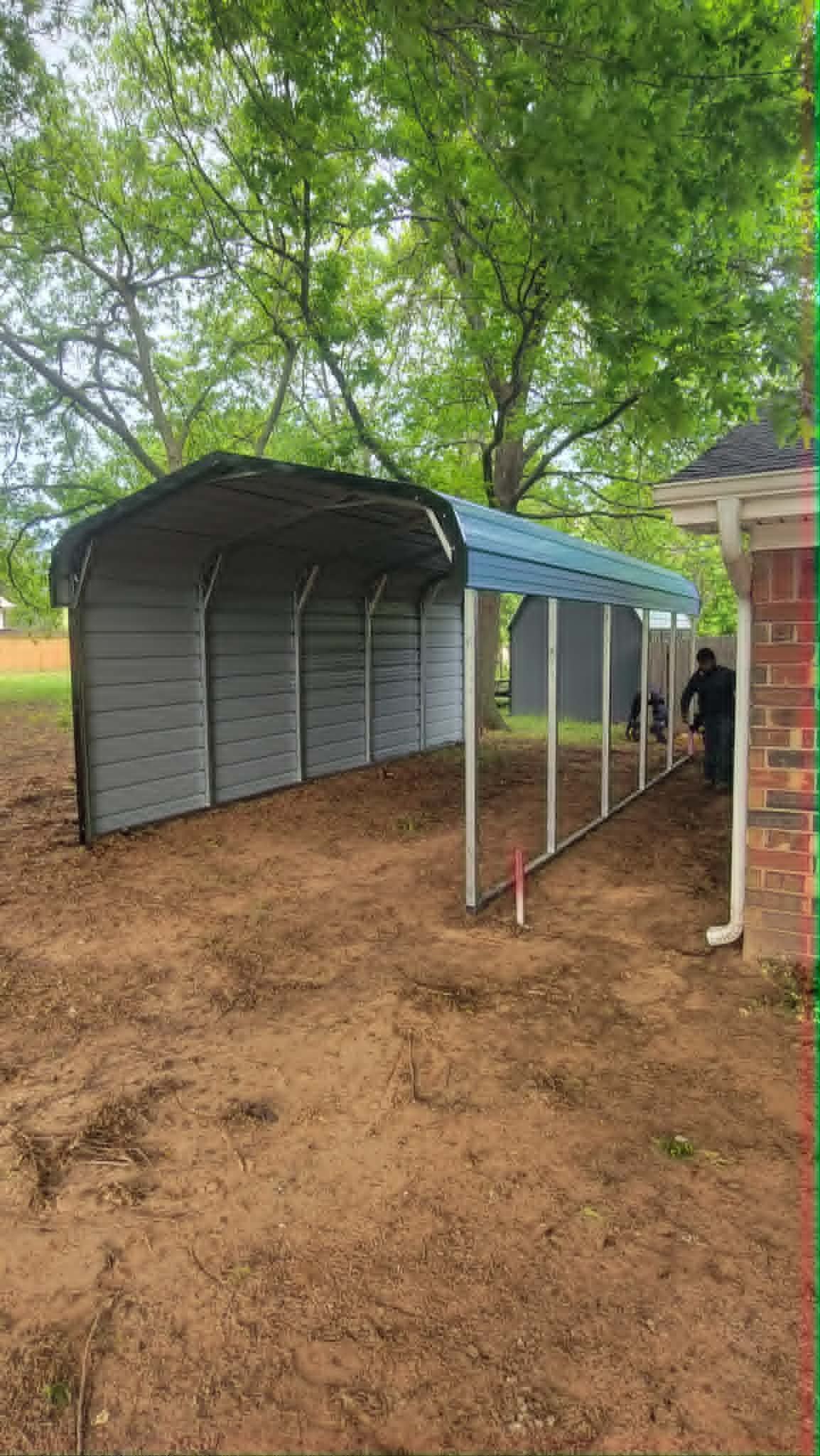 Covered outdoor walkway beside a corrugated shed under trees on a dirt ground
