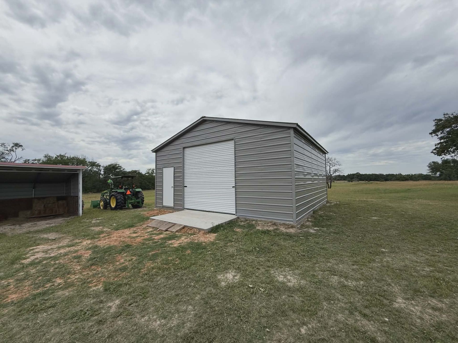 Small gray shed with white double doors on a grassy field under a cloudy sky.