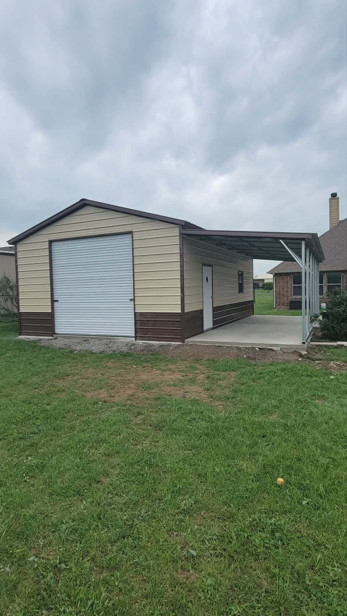Small beige shed with a white door beside a concrete driveway and grassy yard under cloudy sky
