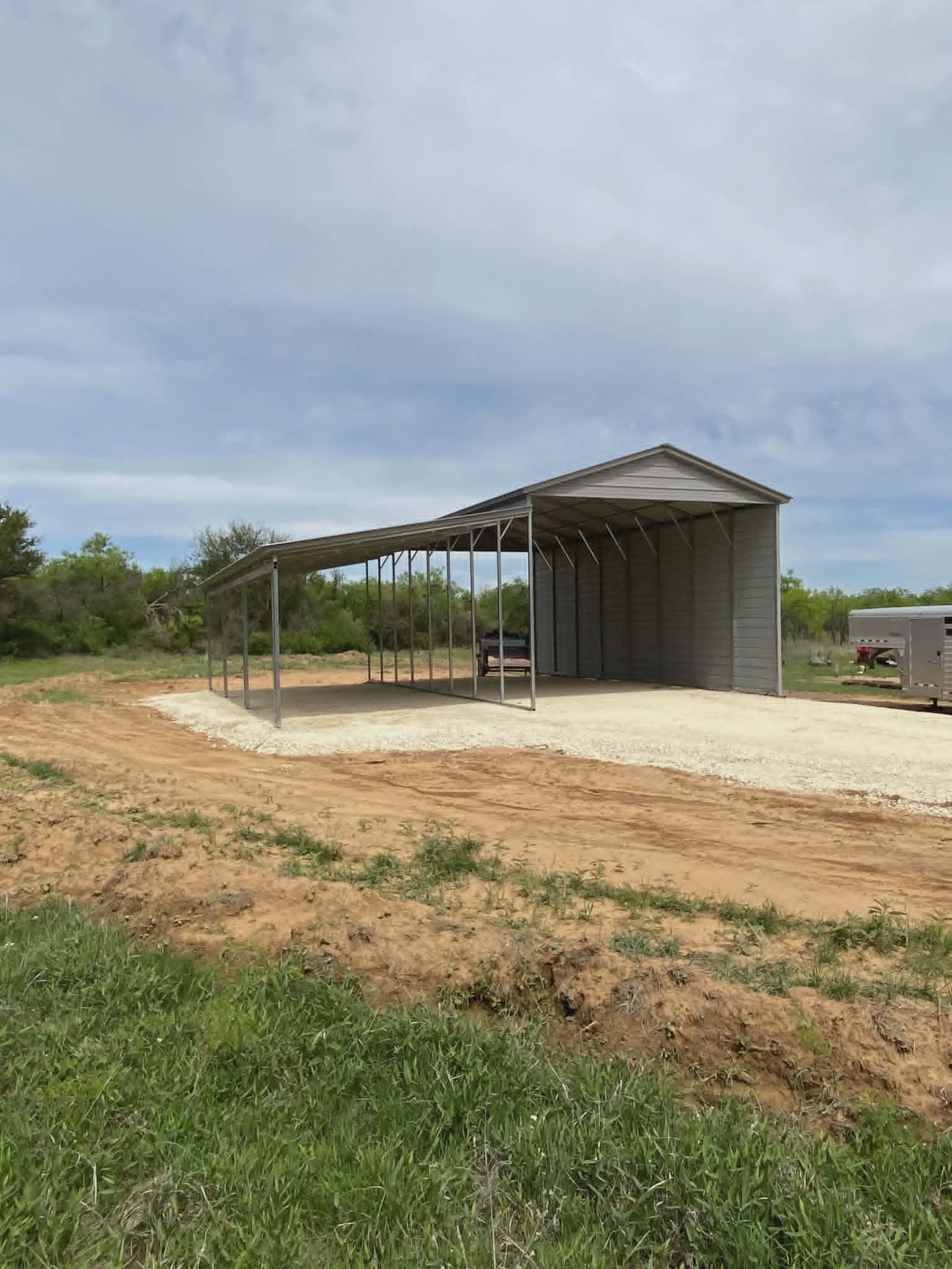 Rural metal shed under construction on a dirt lot, with grassy foreground and cloudy sky.