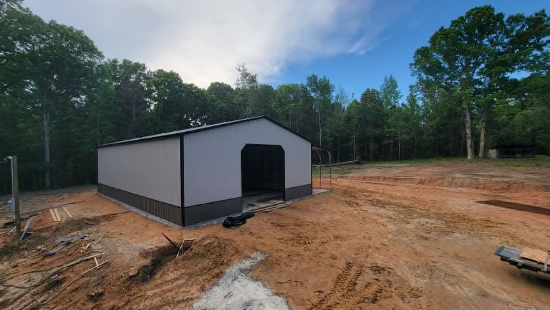 New white-and-black metal shed on a cleared dirt lot beside trees under a partly cloudy sky