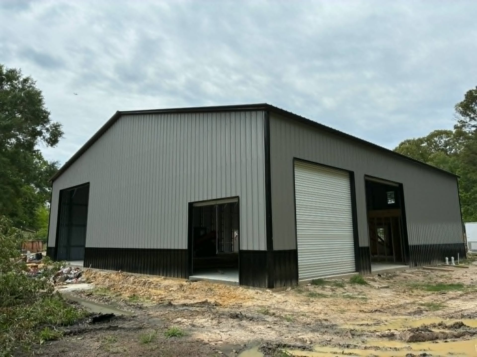 Large gray metal workshop building with black trim, set on a dirt lot under a cloudy sky