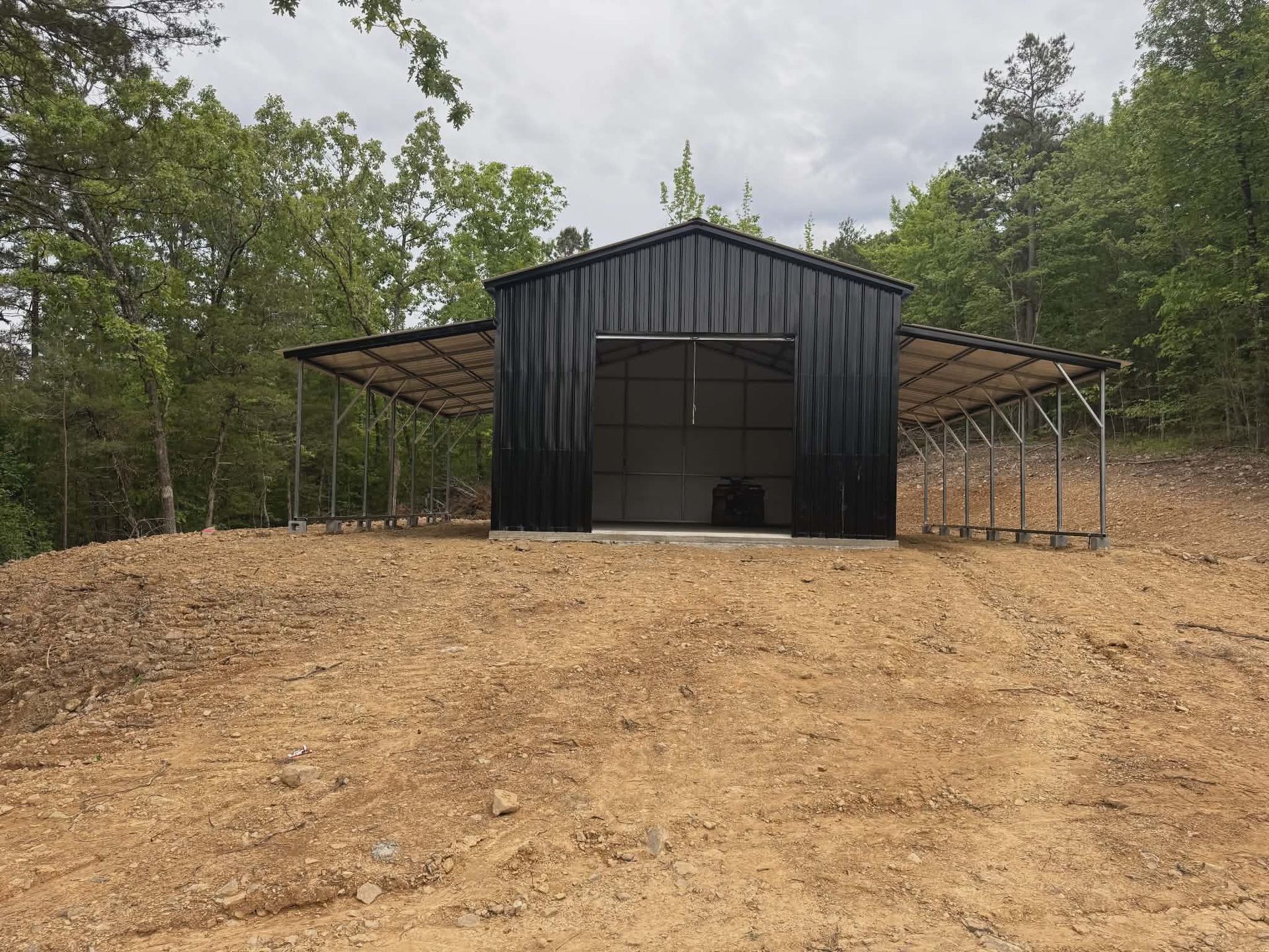 Black metal barn with open sides on a dirt clearing, surrounded by trees under an overcast sky