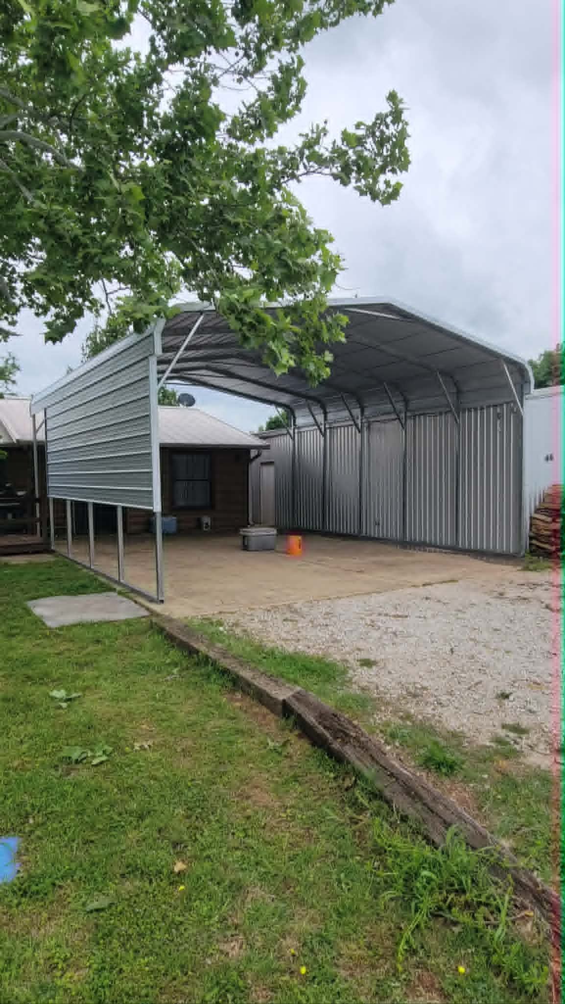 Covered gravel carport beside a grassy yard, with a metal roof and a tree overhead.