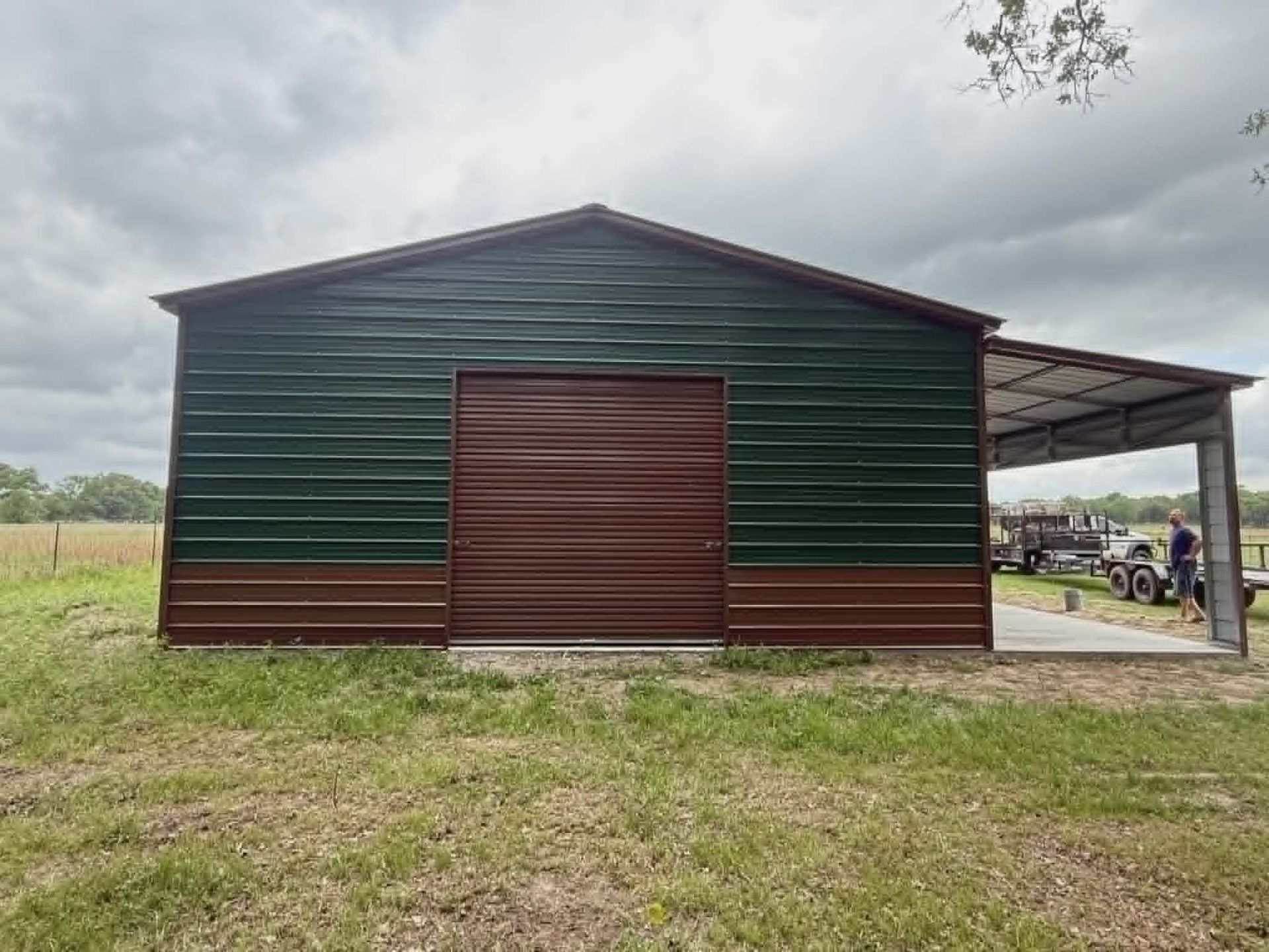 Green and brown metal barn with open side shelter in a grassy field under cloudy sky