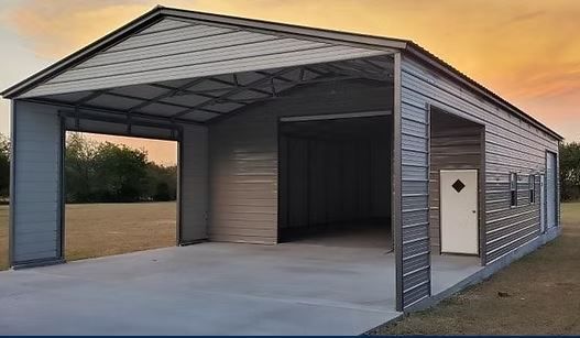 Open-sided metal carport with a concrete floor and a small side door at dusk