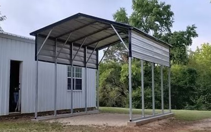 Metal carport attached to a white building, with a slanted roof and open sides on a grassy lot.