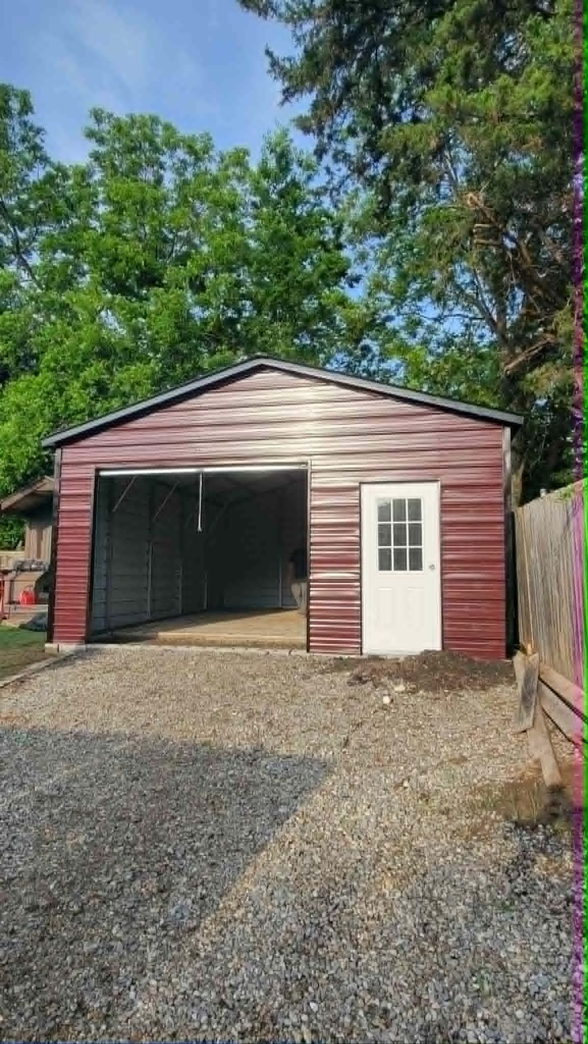 Red wooden garage with open door, white side door, and gravel driveway surrounded by trees.