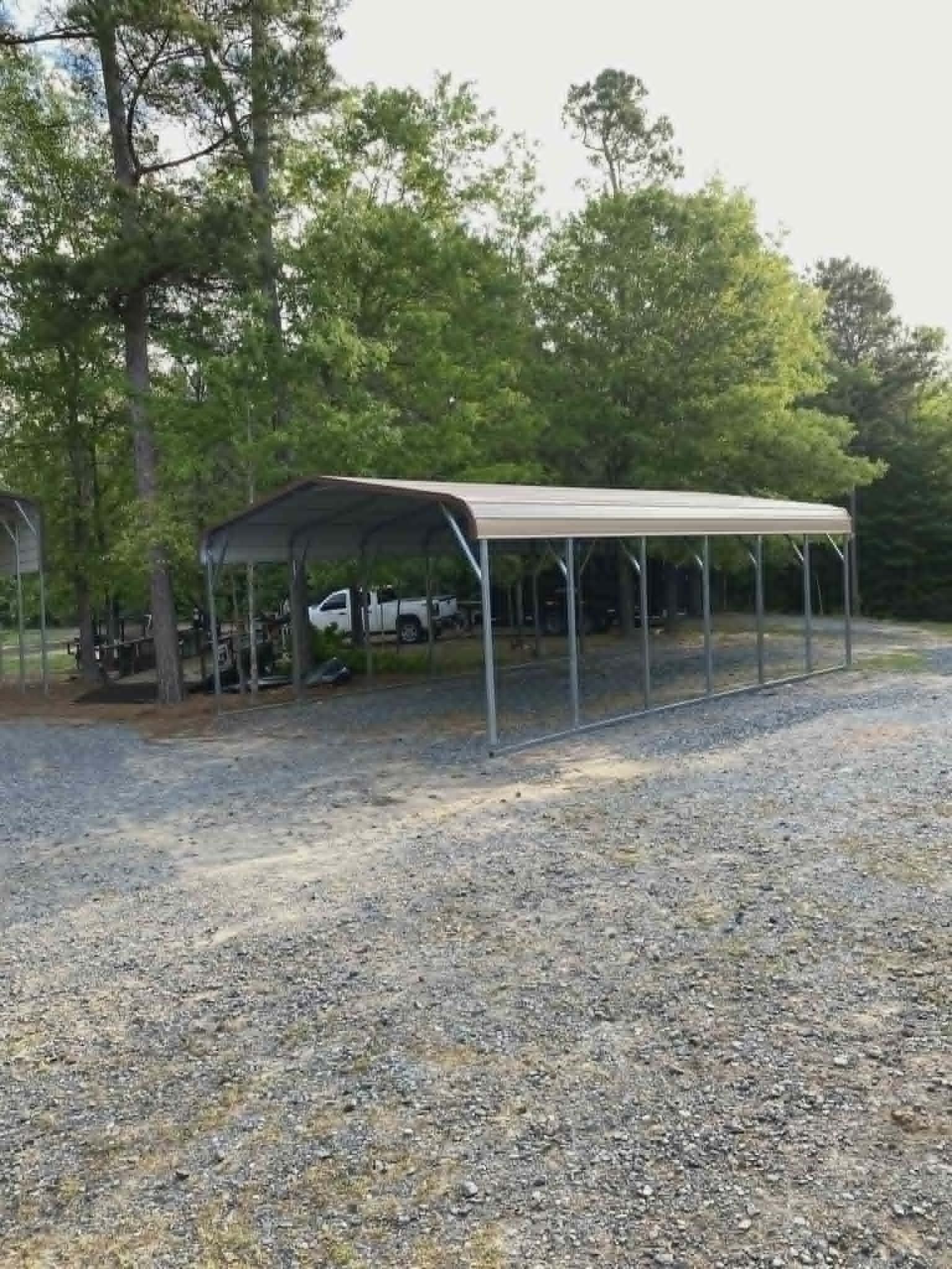 Gravel lot with a metal carport sheltering vehicles beside trees in a wooded area
