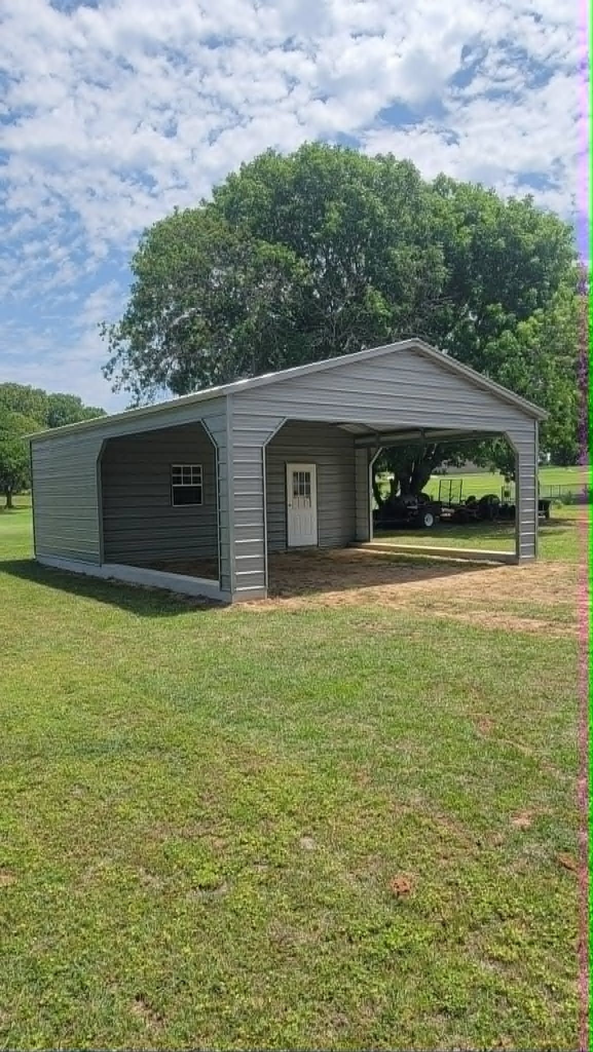 Small white open-sided shed on a grassy field under a cloudy sky, with a large tree behind it