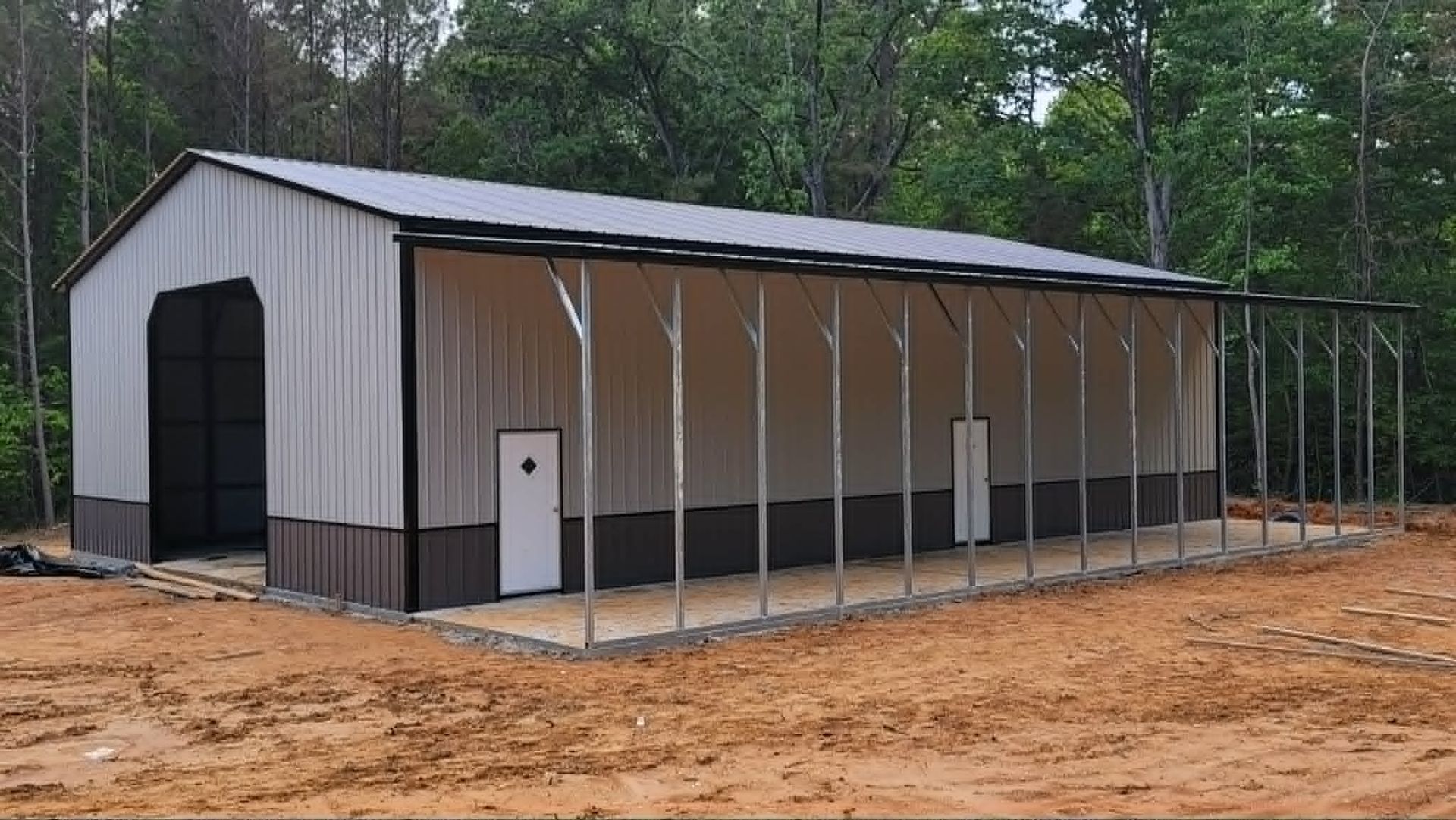 Large gray metal building with open-sided porch on a dirt lot beside trees