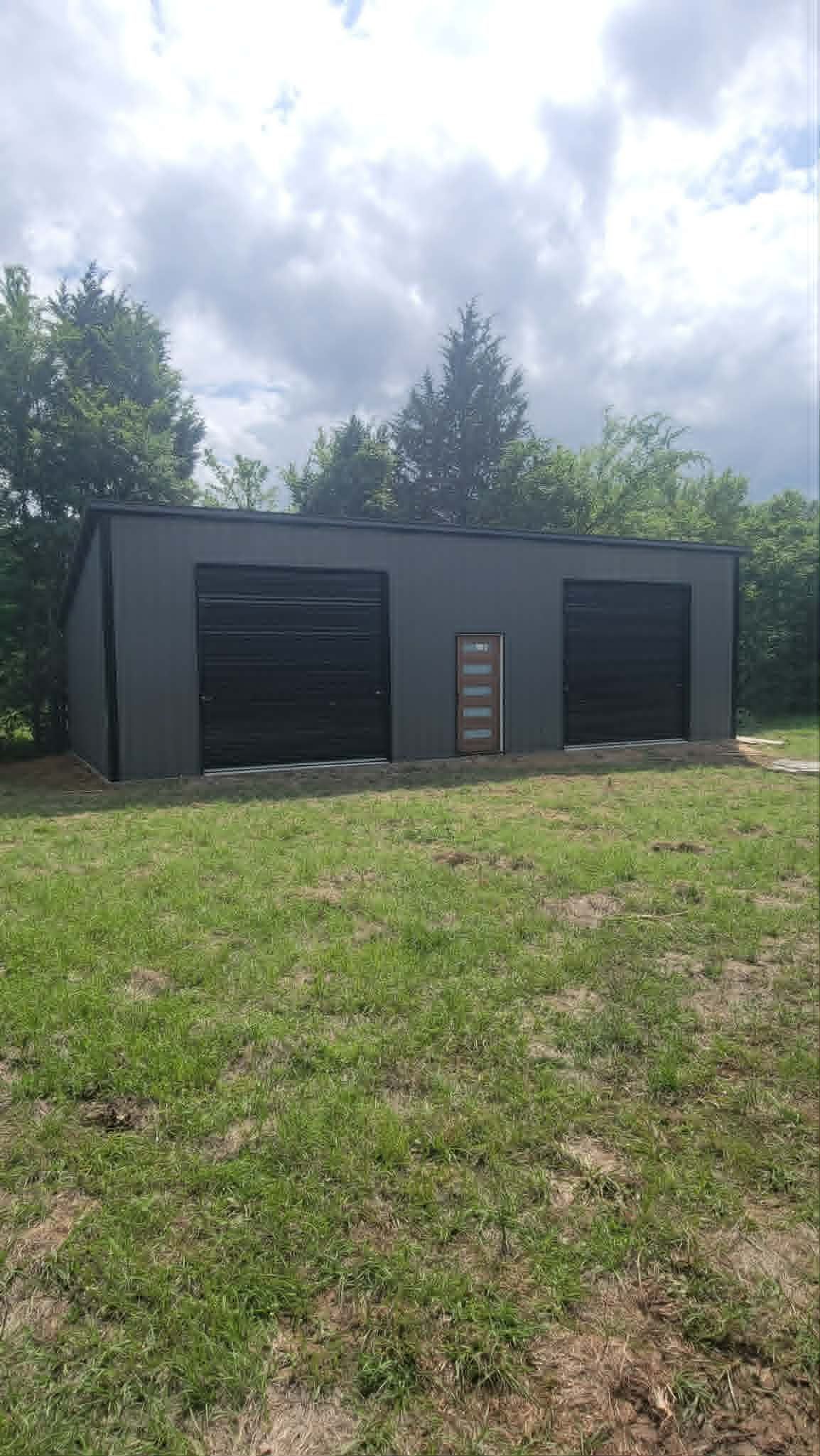 Small gray outbuilding with two dark doors and a ladder, set in a grassy yard under cloudy sky