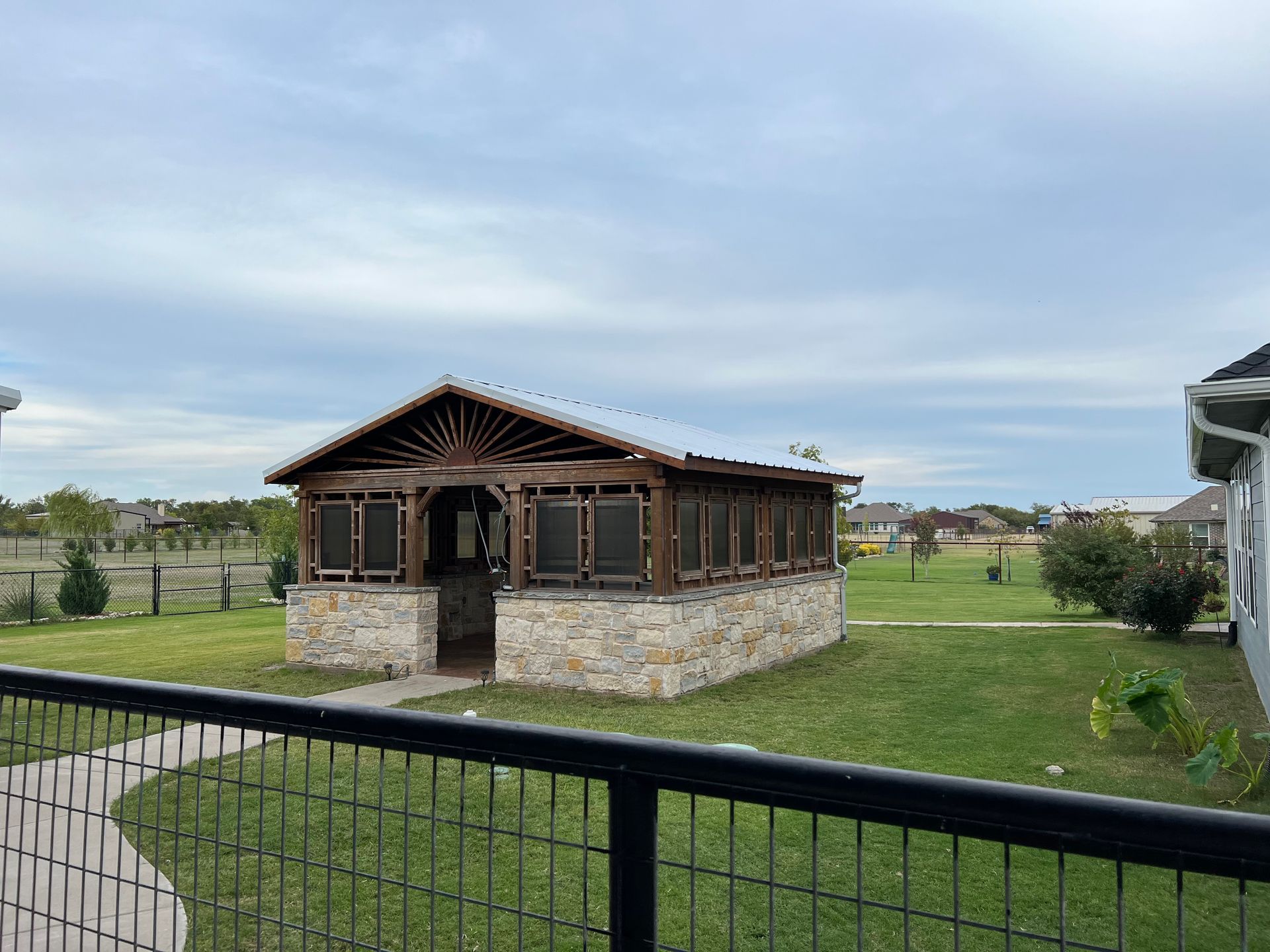 Small wooden cabin with stone base in a grassy yard, viewed behind a black metal fence under a cloudy sky