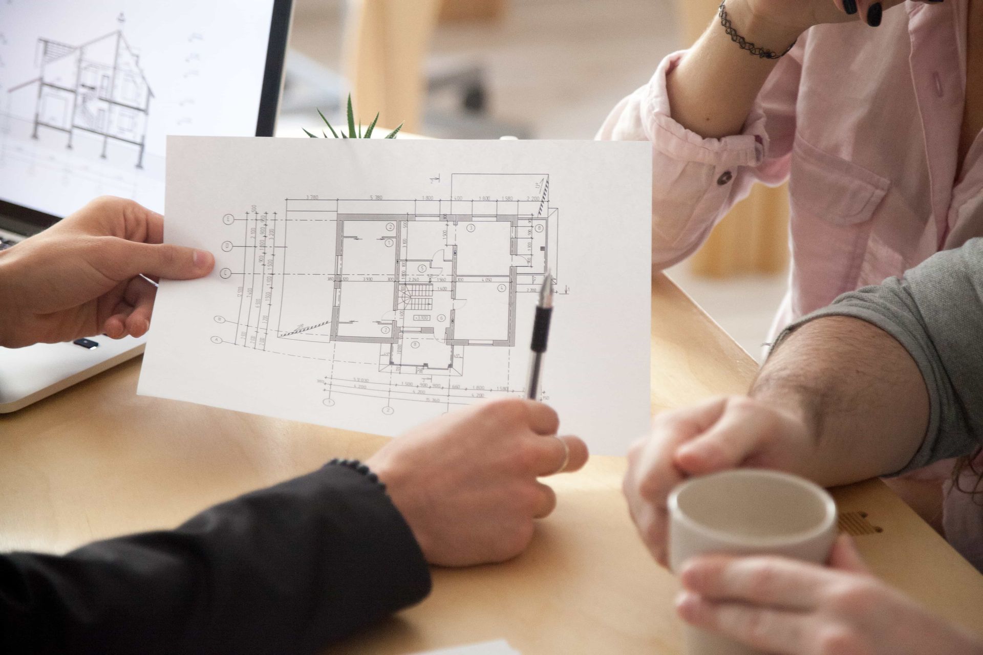 Hands discussing an architectural floor plan at a table, with coffee cups and sketch papers visible.