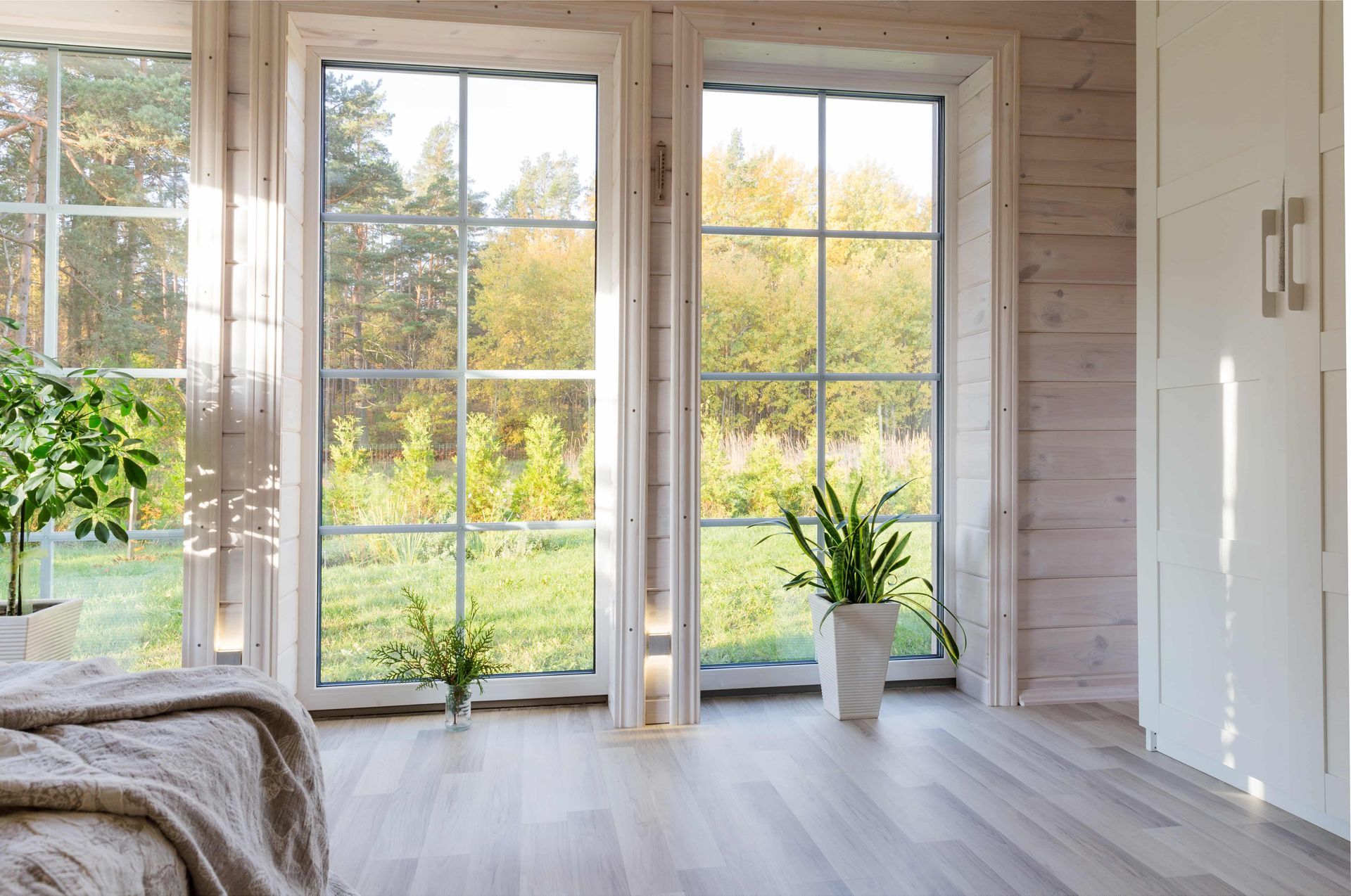 Bright living room with large windows, light wood floors, and plants overlooking a green garden