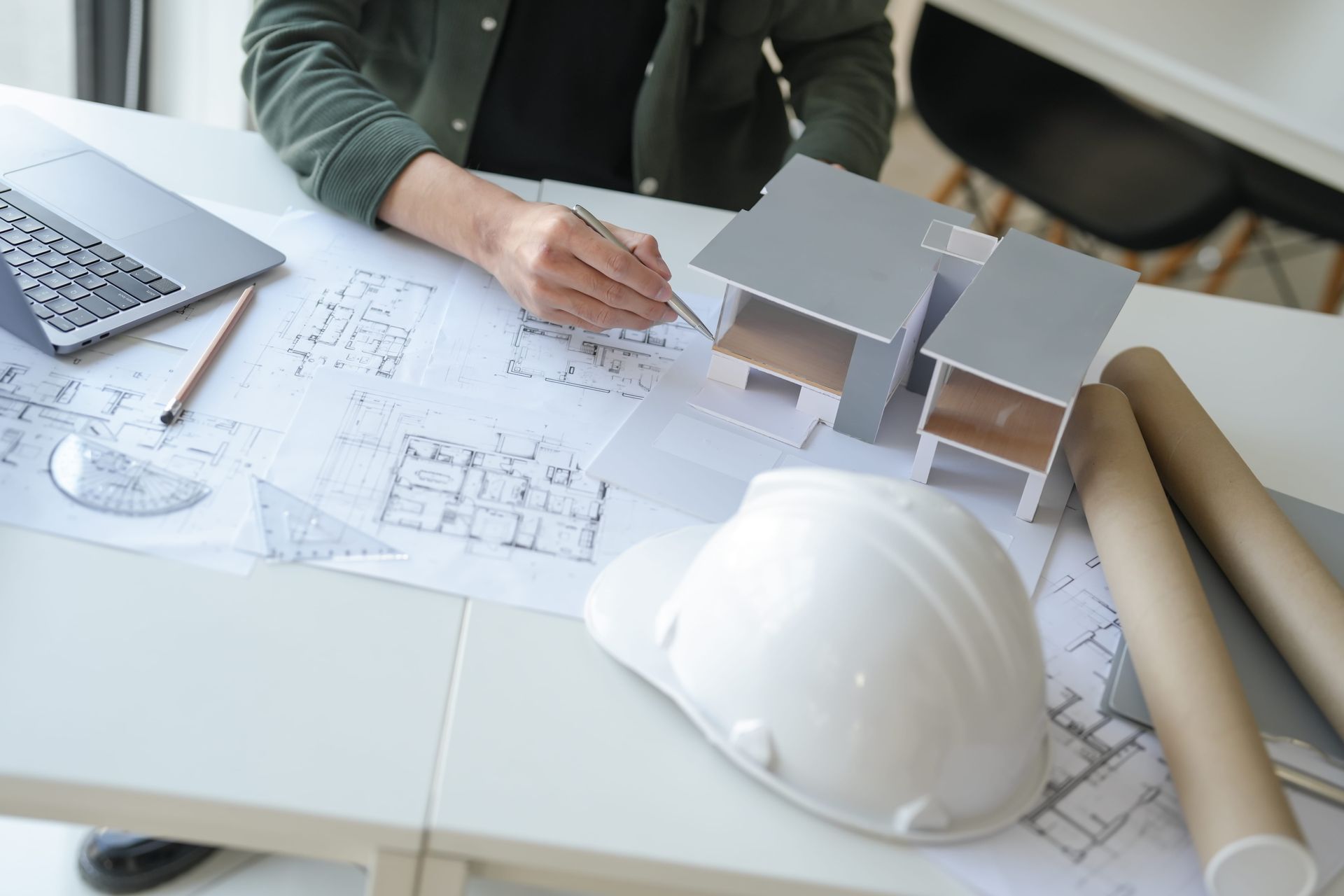 Person sketching building plans at a desk with blueprints, laptop, model houses, and a white hard hat