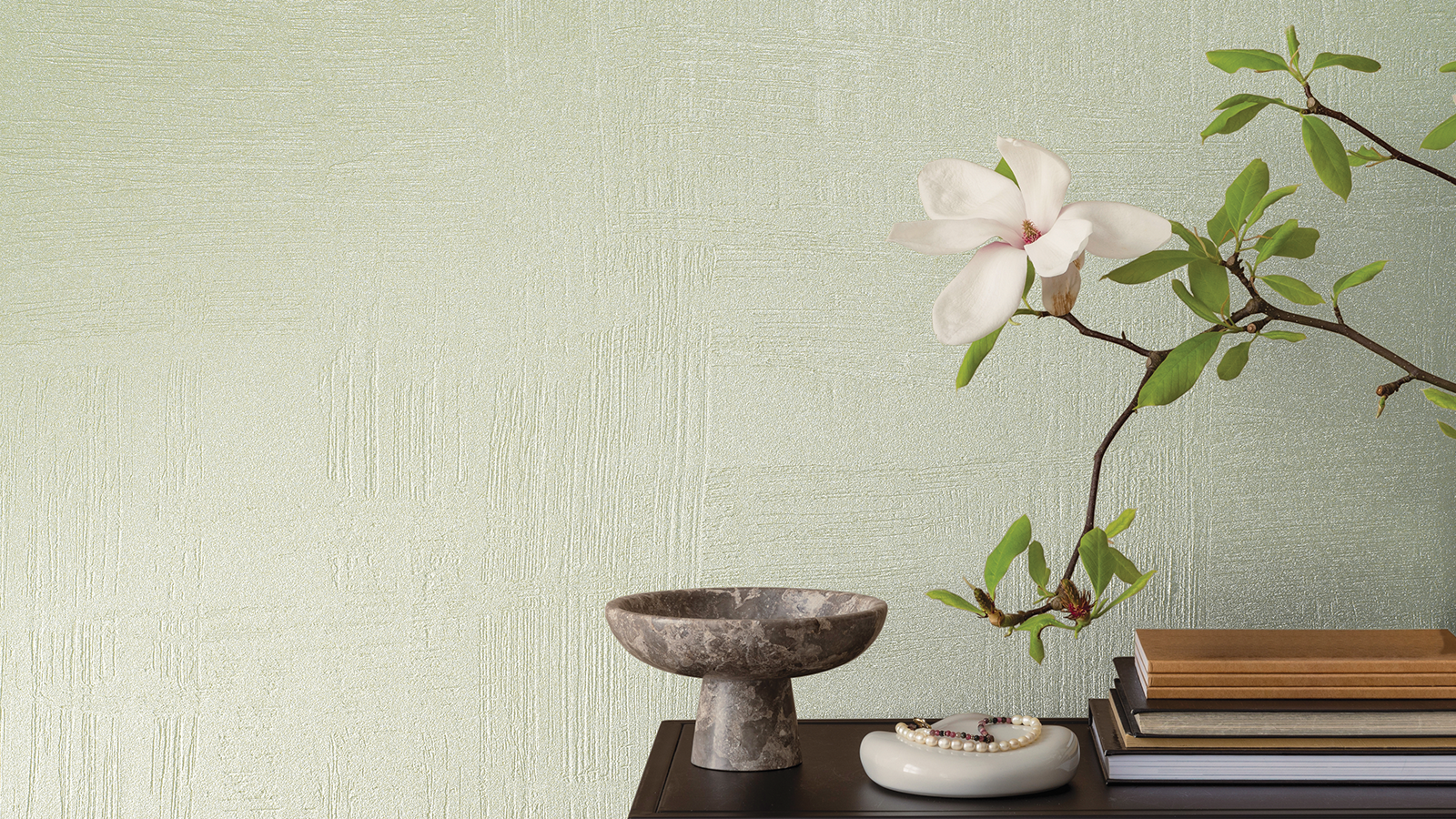 Uno sfondo da parete con texture verde chiaro, un ramo di magnolia, libri e una ciotola decorativa su un tavolo scuro.