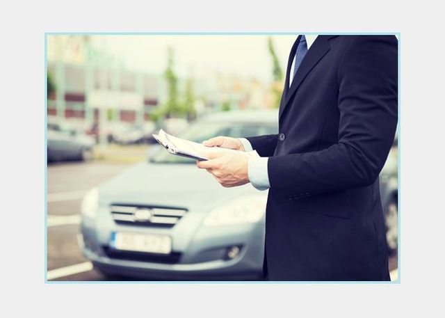 Hombre con traje oscuro sosteniendo documentos junto a un coche plateado al aire libre