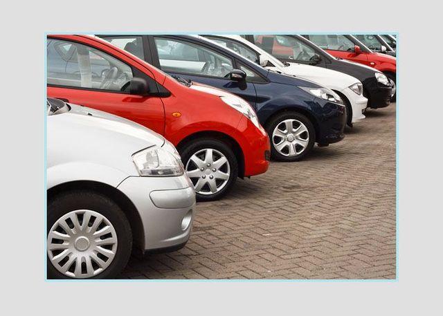 Fila de coches aparcados en un estacionamiento, incluyendo vehículos plateados, rojos, negros y blancos.