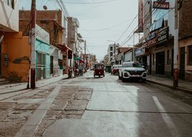 Calle tranquila con tiendas, coches aparcados y un vehículo de tres ruedas bajo cables eléctricos.